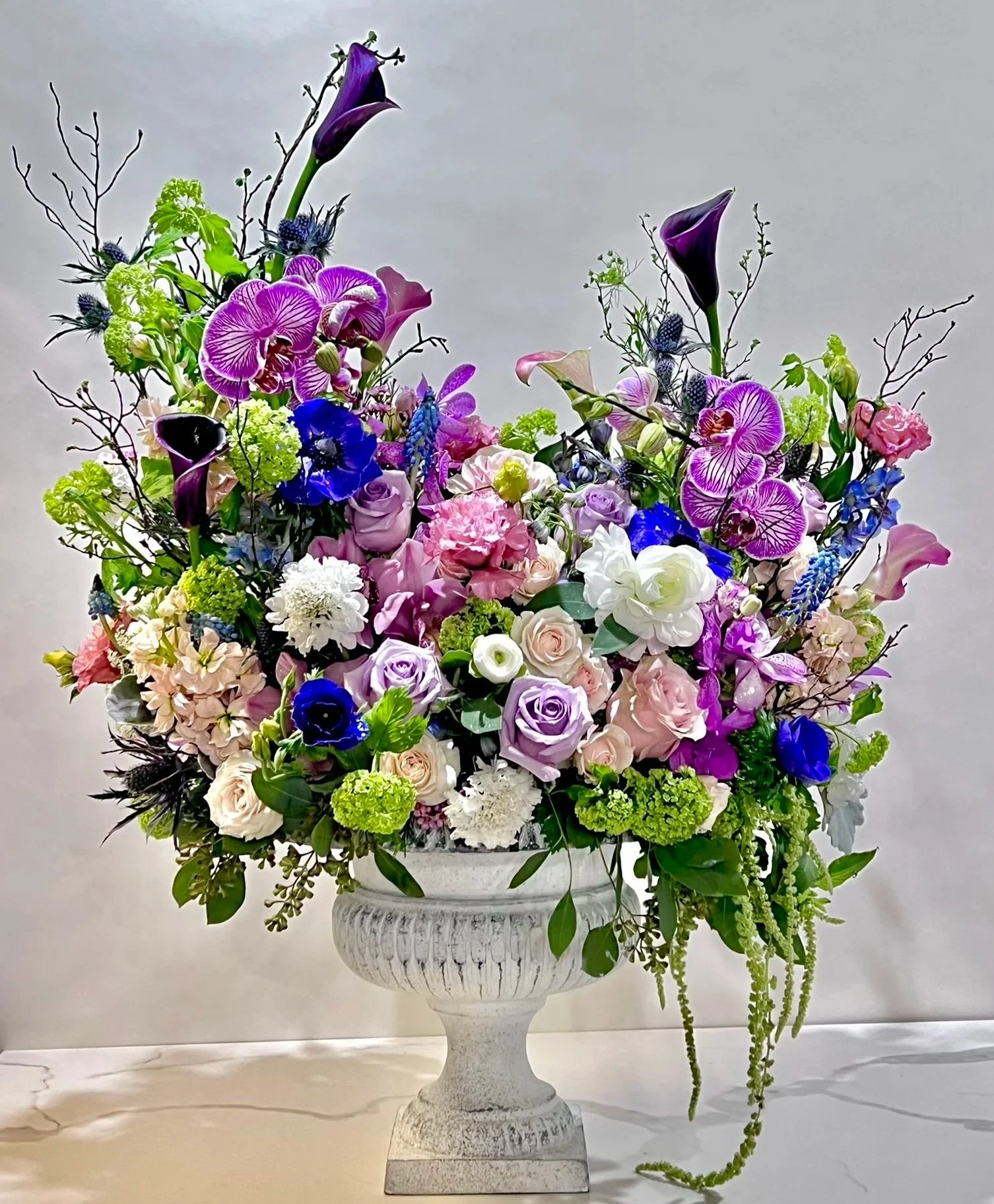 A large, elegant floral arrangement in a white pedestal vase with pink, purple, blue, green, and white flowers, including orchids, roses, and calla lilies, along with greenery and decorative twigs, set against a plain background.