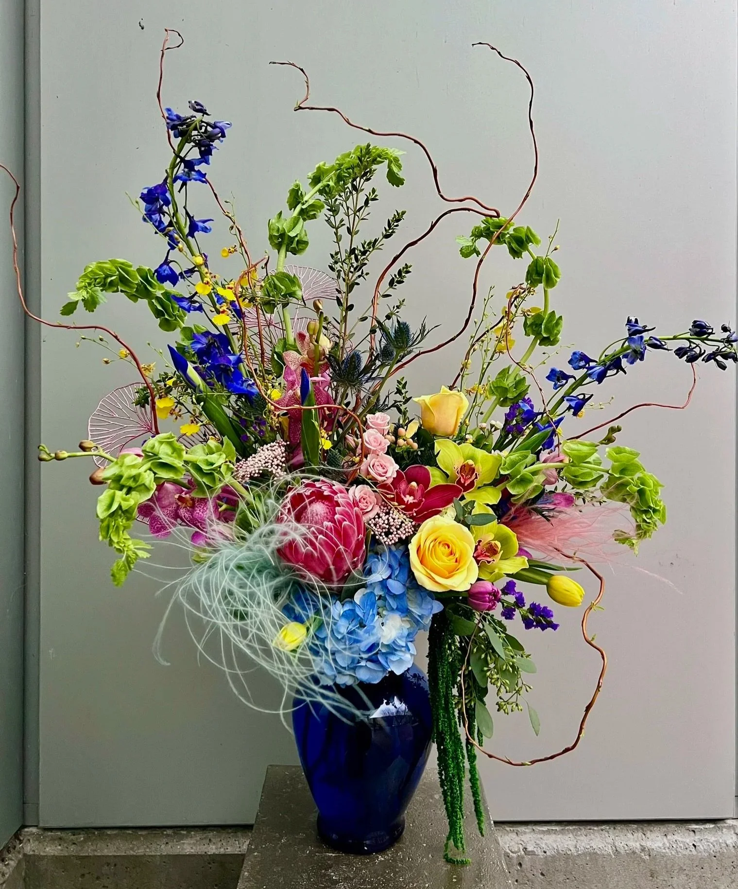 A colorful floral arrangement in a dark blue vase featuring blue, pink, yellow, and purple flowers, with curly branches and white decorative mesh.