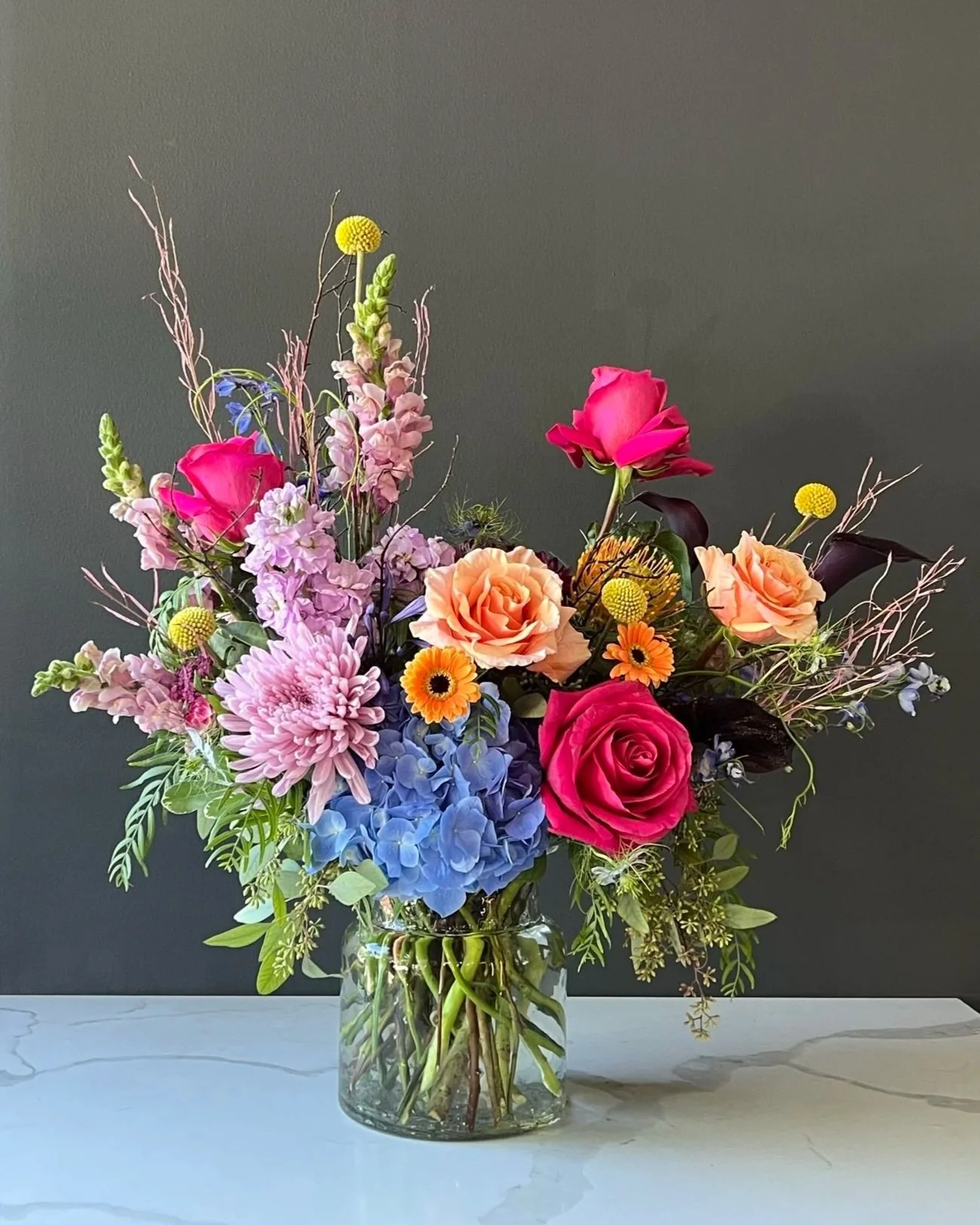 A colorful flower bouquet with pink roses, orange and blue hydrangeas, yellow billy balls, and various greenery in a clear glass vase on a white surface against a dark background.