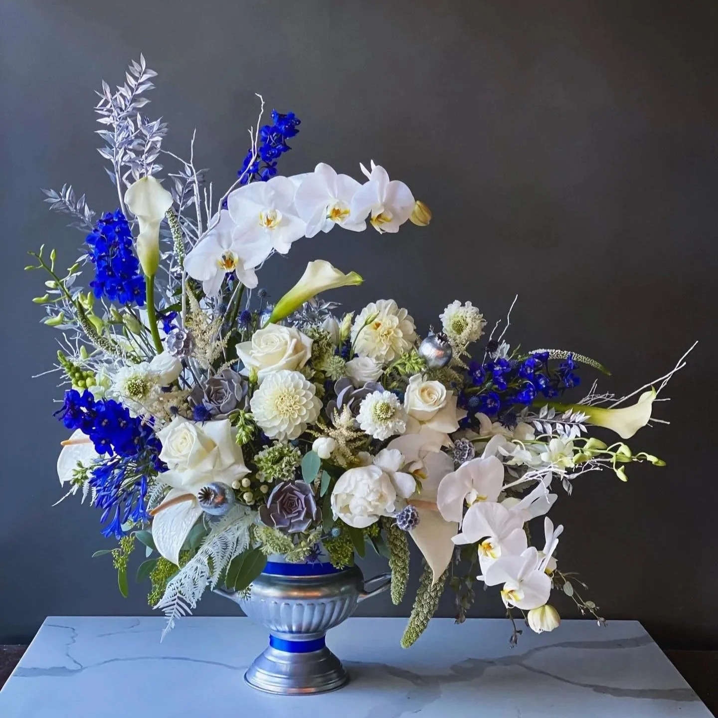 Elegant flower arrangement in a silver and blue vase featuring white roses, orchids, calla lilies, and blue delphiniums on a white marble surface against a dark background.