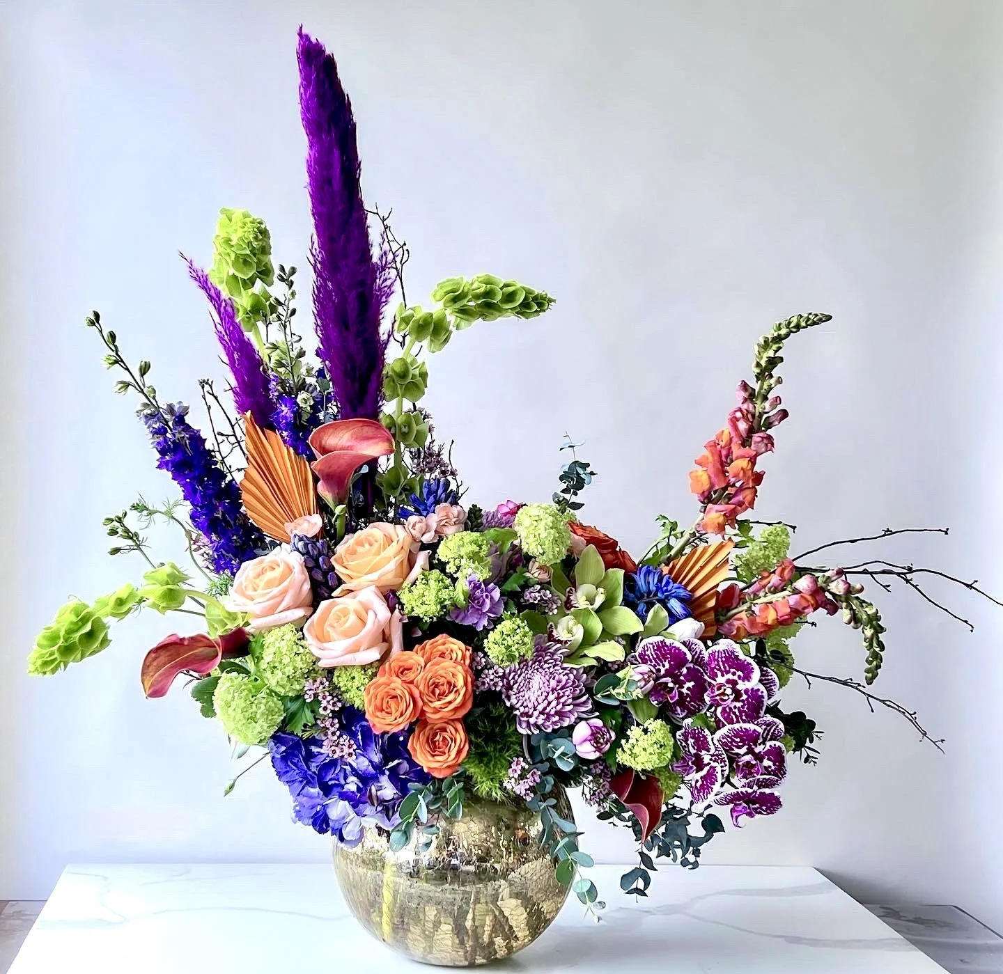 Colorful floral arrangement in a gold vase featuring roses, orchids, calla lilies, delphiniums, snapdragons, and other flowers against a neutral background.