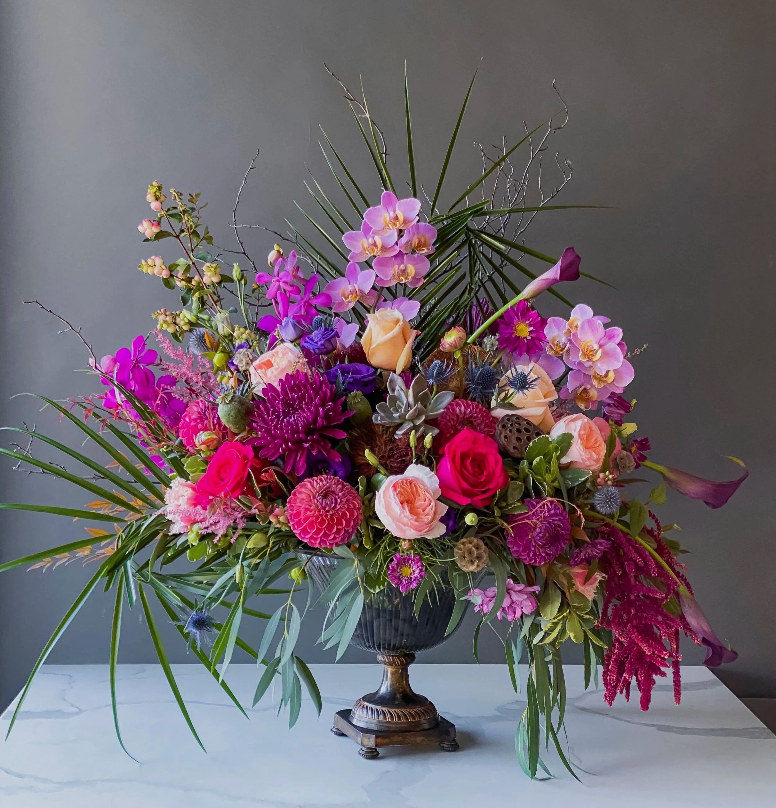 A large floral arrangement in a bronze pedestal vase featuring pink, purple, orange, and red flowers, with greenery and branches against a plain gray background.