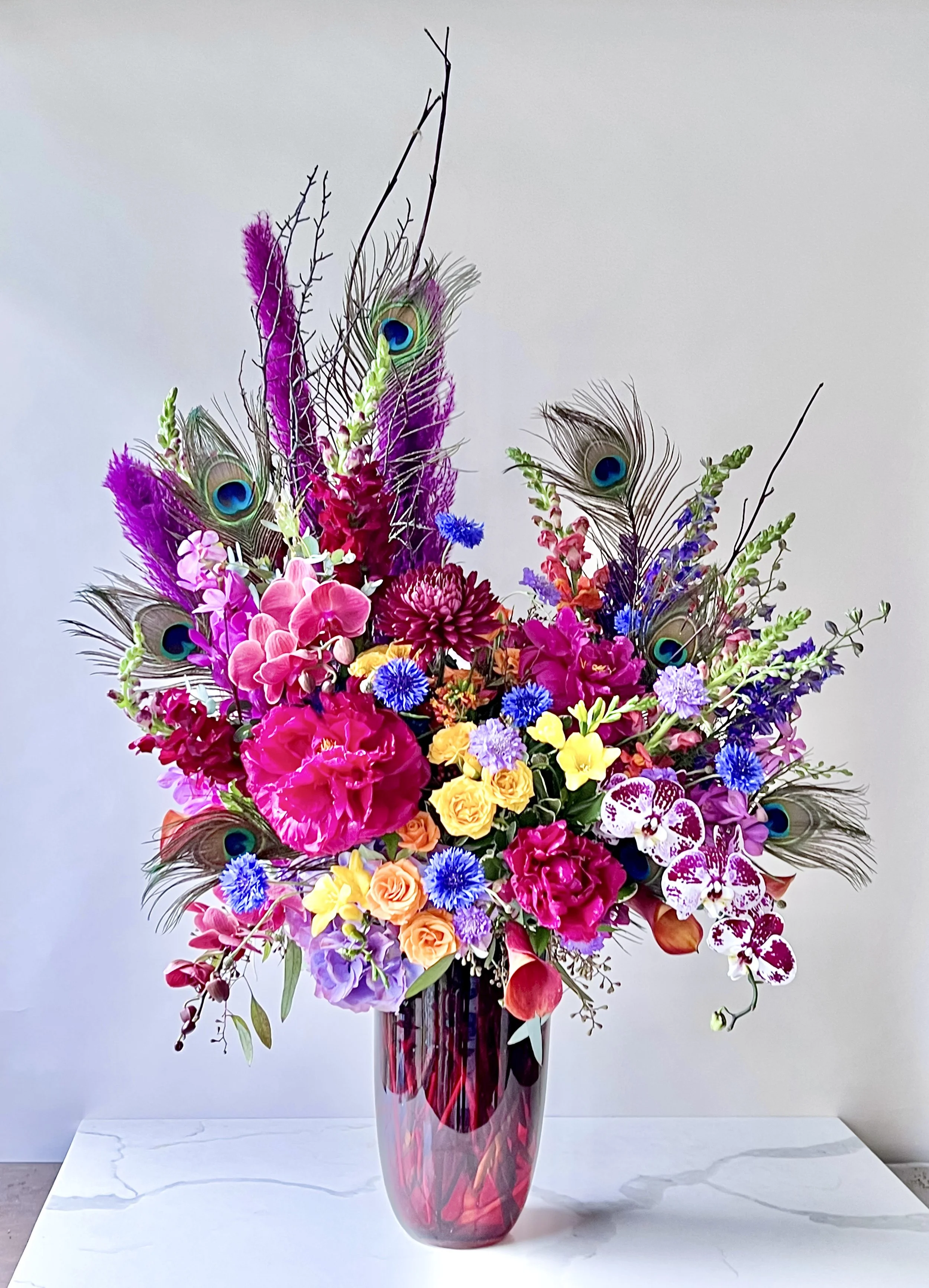 A tall, multicolored flower arrangement in a pink and purple glass vase, featuring pink orchids, purple roses, yellow roses, blue cornflowers, peacock feathers, and various greenery, set against a plain gray wall.