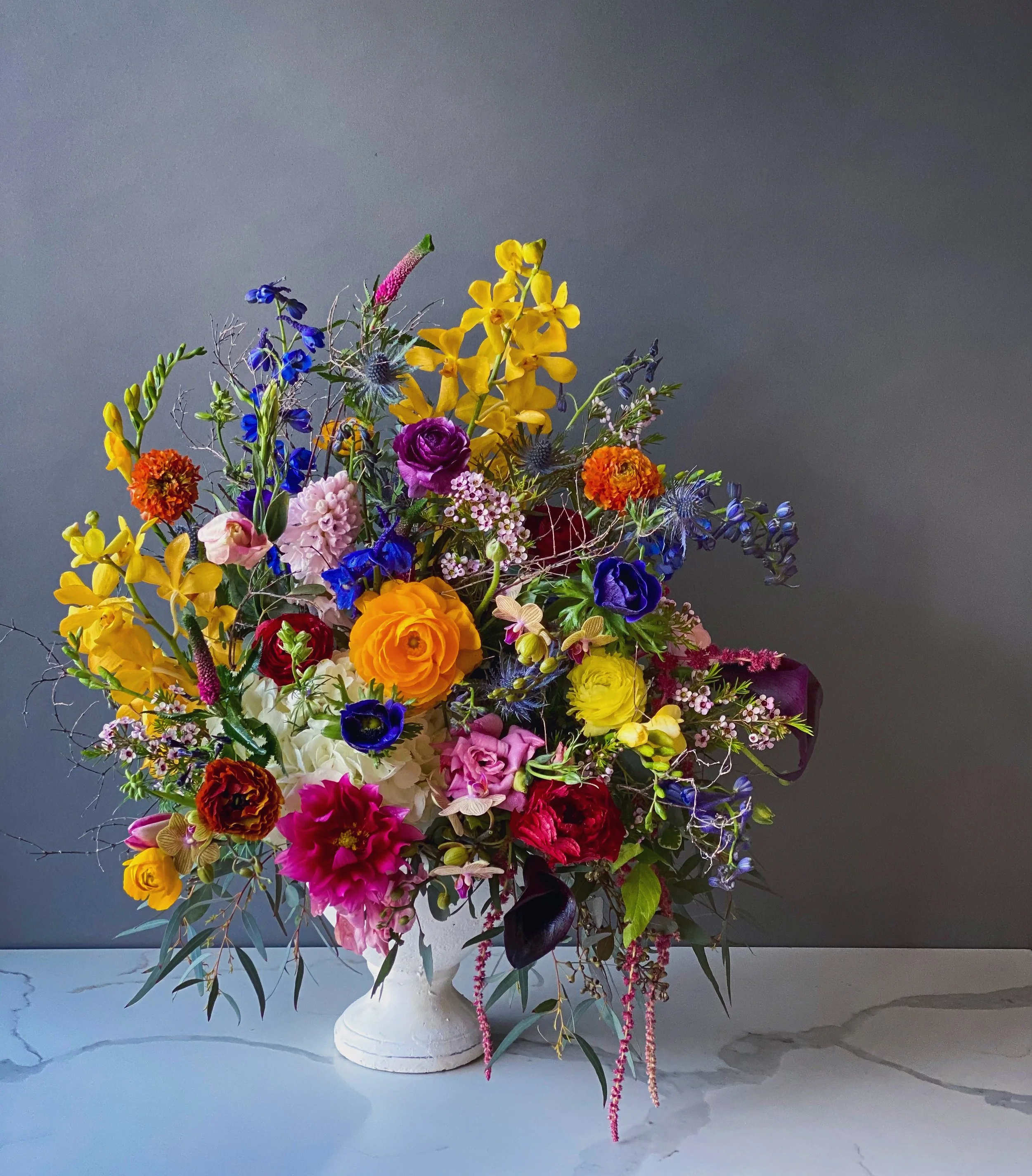 Colorful bouquet of various flowers in a white vase against a plain gray background.