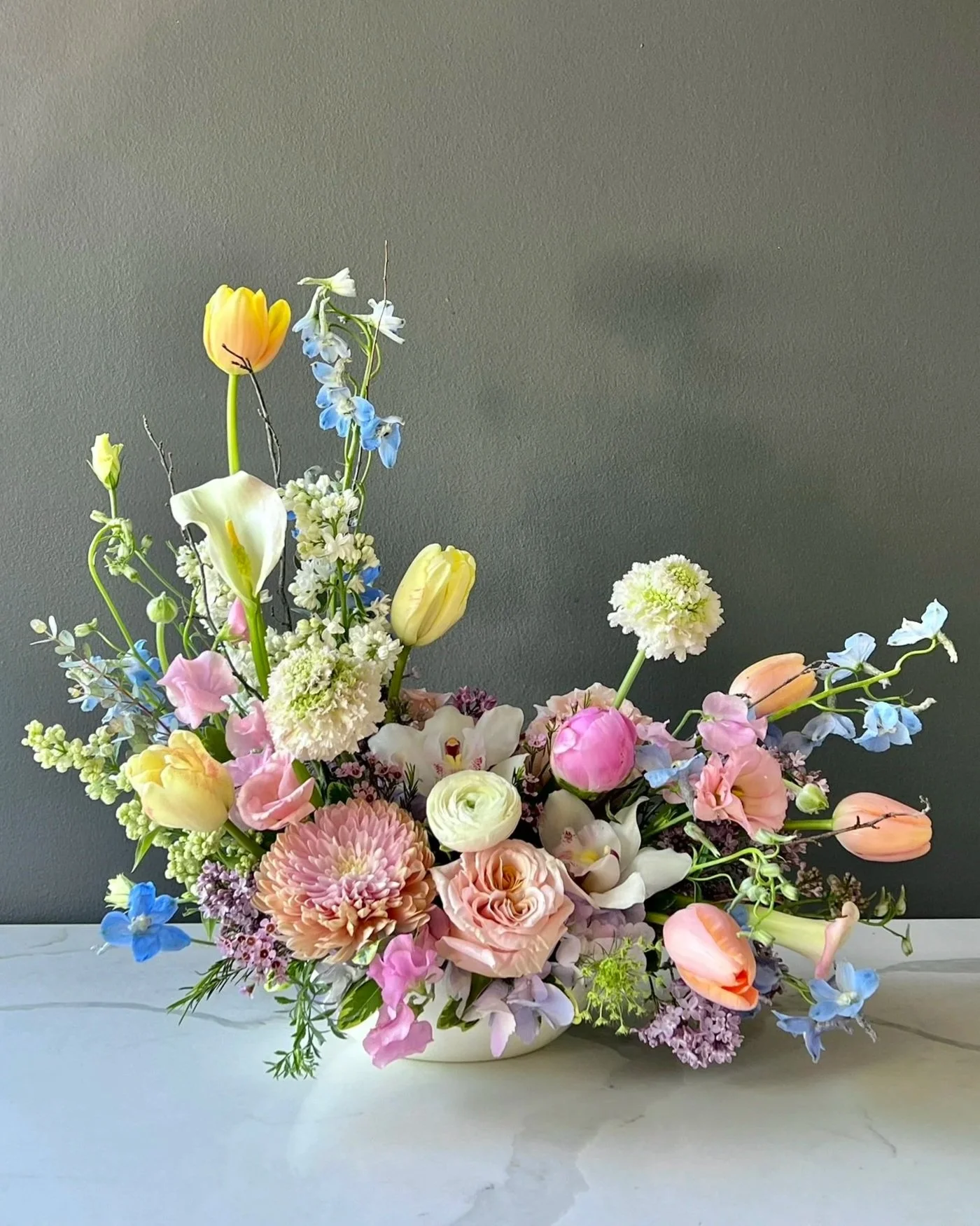 A colorful bouquet of various flowers including tulips, roses, and other blossoms, arranged in a white vase against a gray wall.