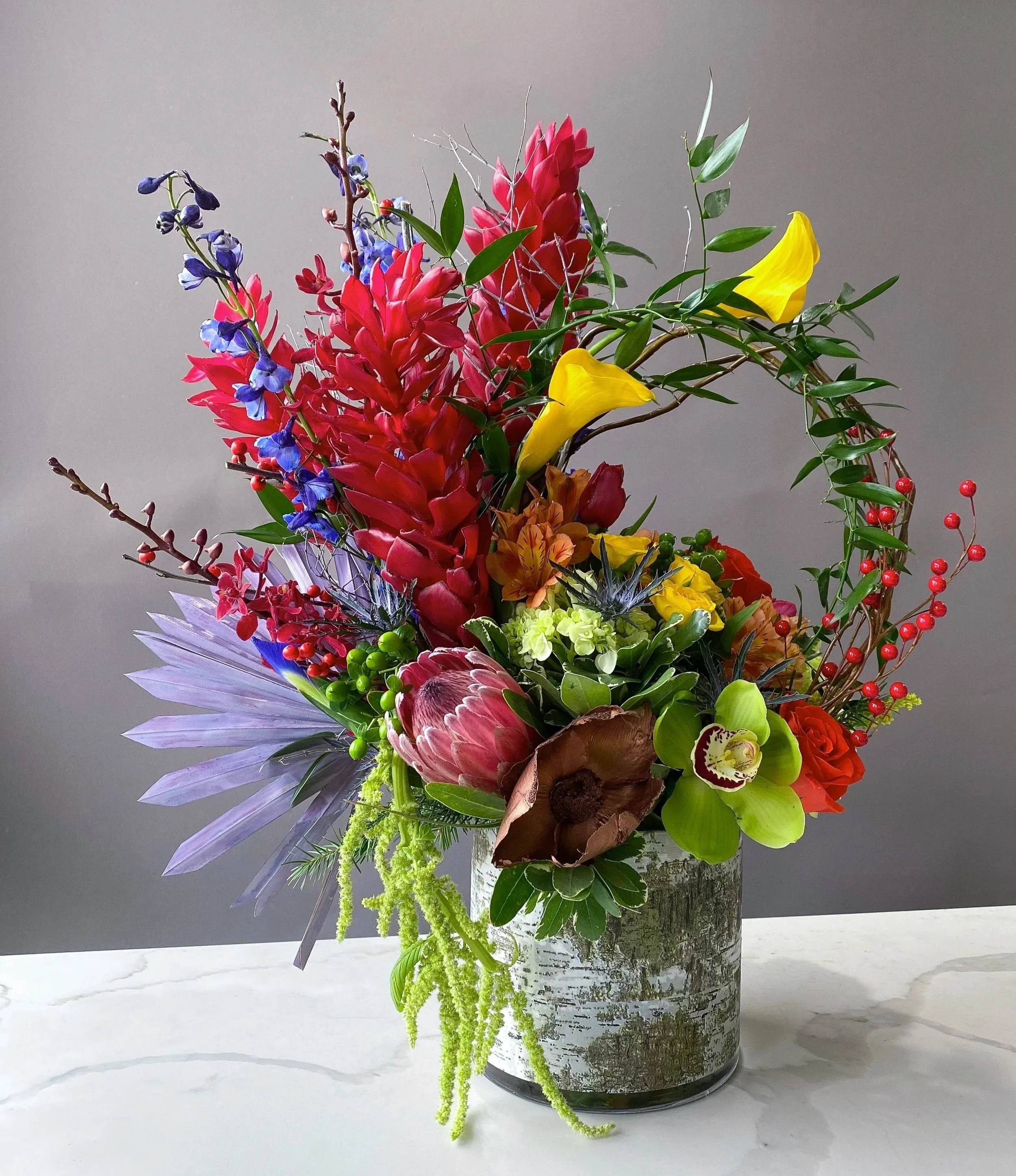 Colorful flower arrangement featuring red, yellow, purple, and green flowers and foliage in a silver vase against a neutral background.