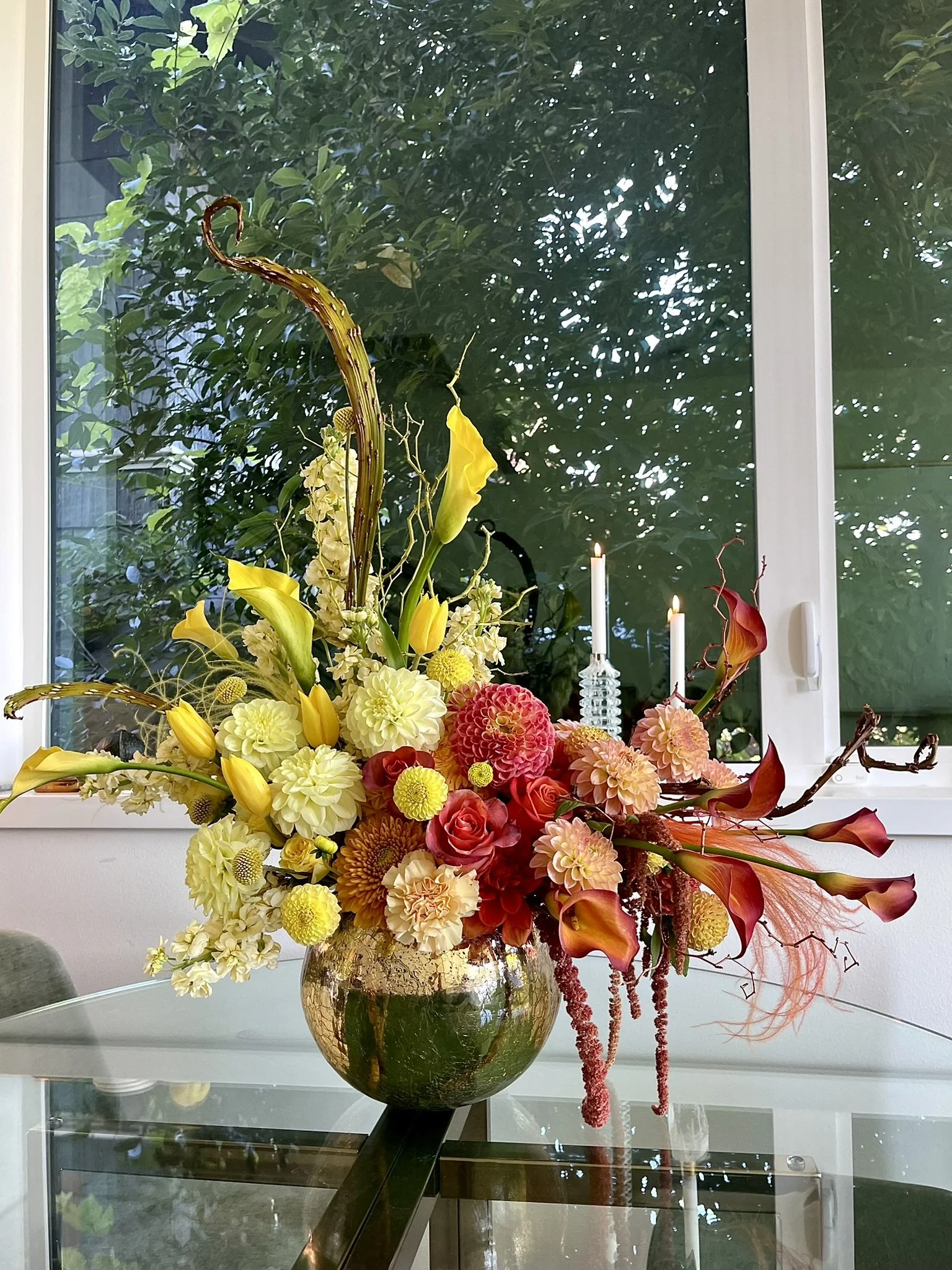 A large floral arrangement in a round, gold-colored vase on a glass table indoors. The bouquet contains yellow lilies, white and yellow dahlias, red roses, and orange calla lilies, with long red and pink hanging amaranthus. There are two lit white candles in glass holders behind the flowers. A window with greenery outside is in the background.