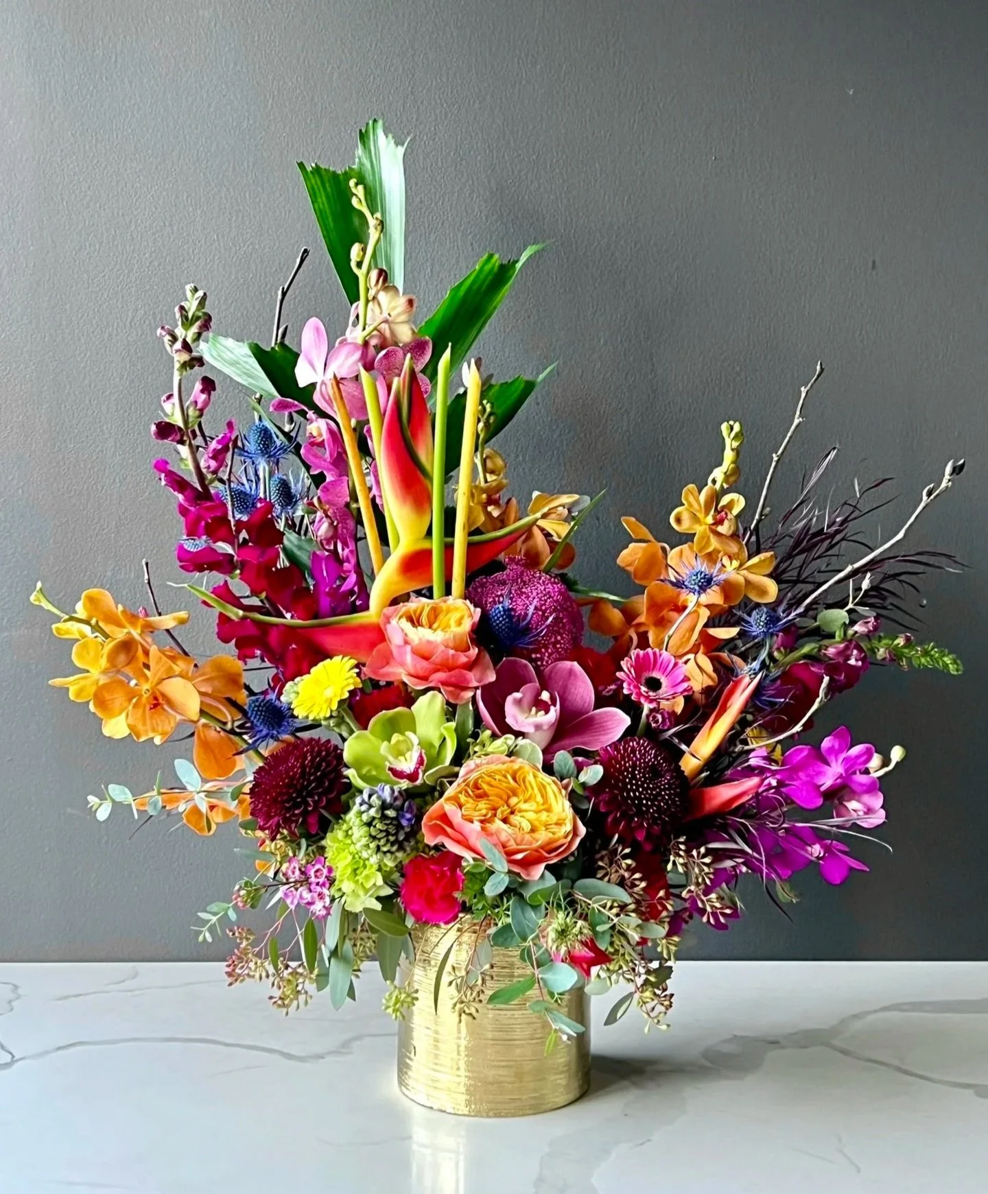 Colorful floral arrangement in a gold vase on a white marble surface against a gray background.