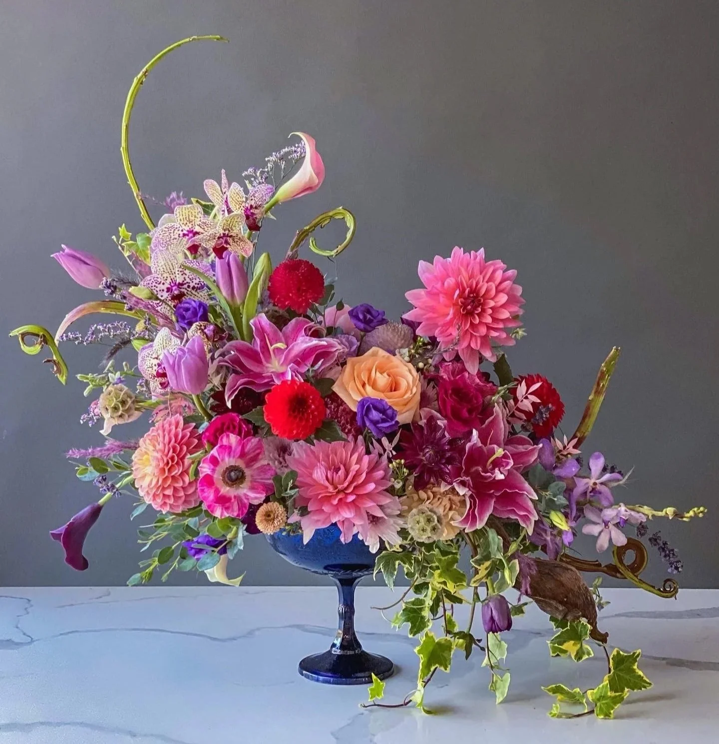 A colorful flower arrangement with pink, purple, red, and peach flowers in a blue glass vase on a white surface against a plain gray background.