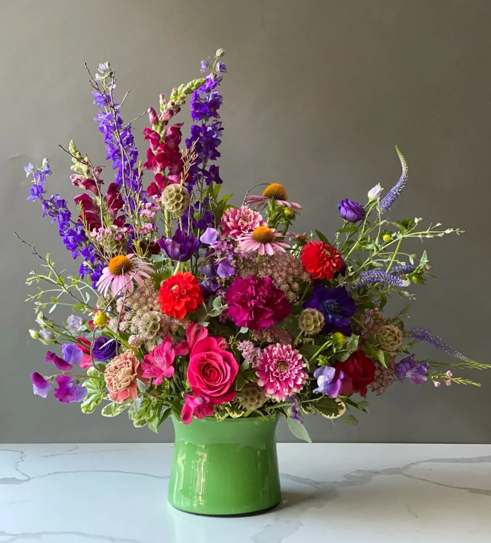 A large bouquet of colorful flowers in a green vase on a marble surface with a plain background.