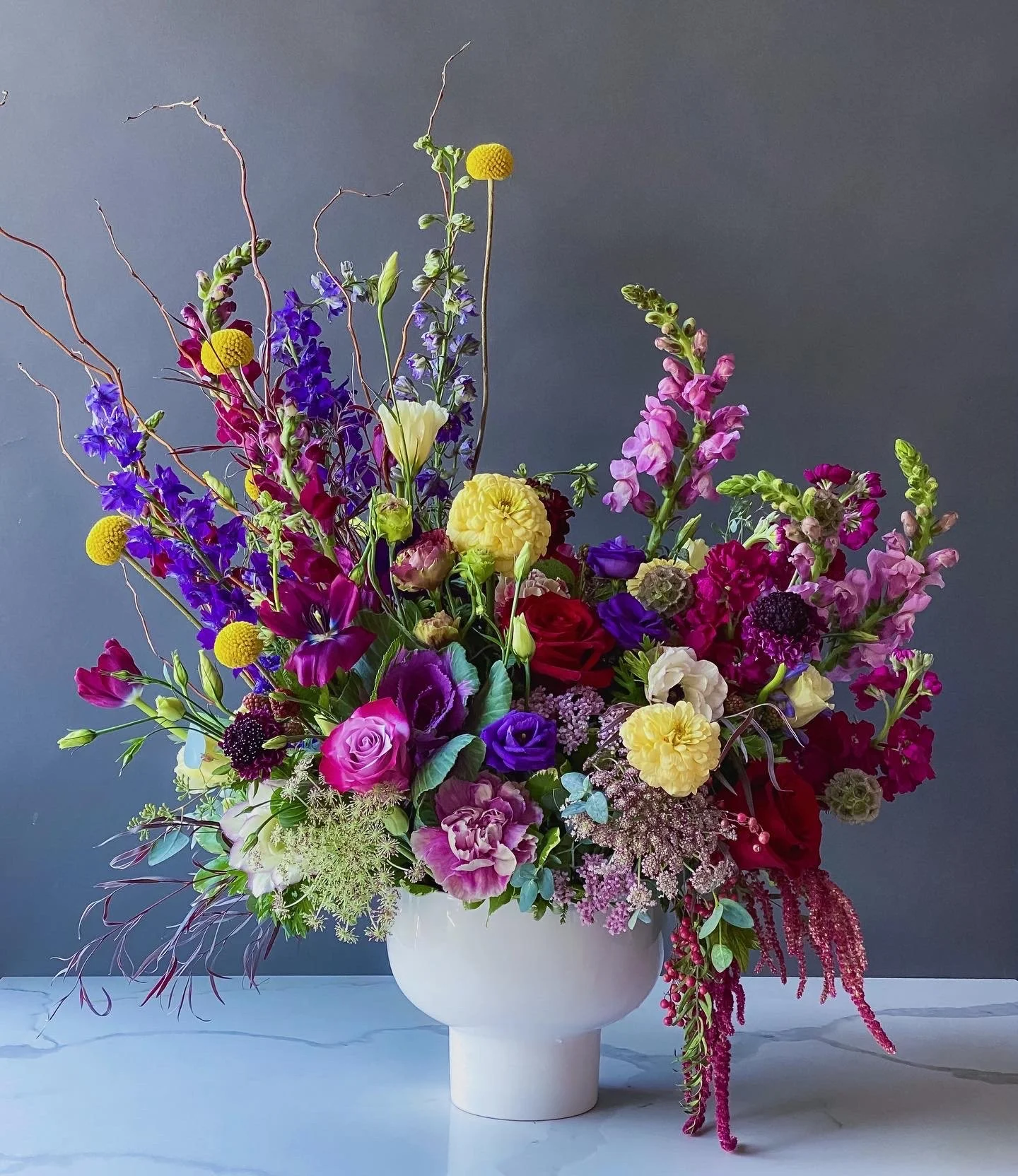 A colorful bouquet of various flowers in a white vase against a gray background.