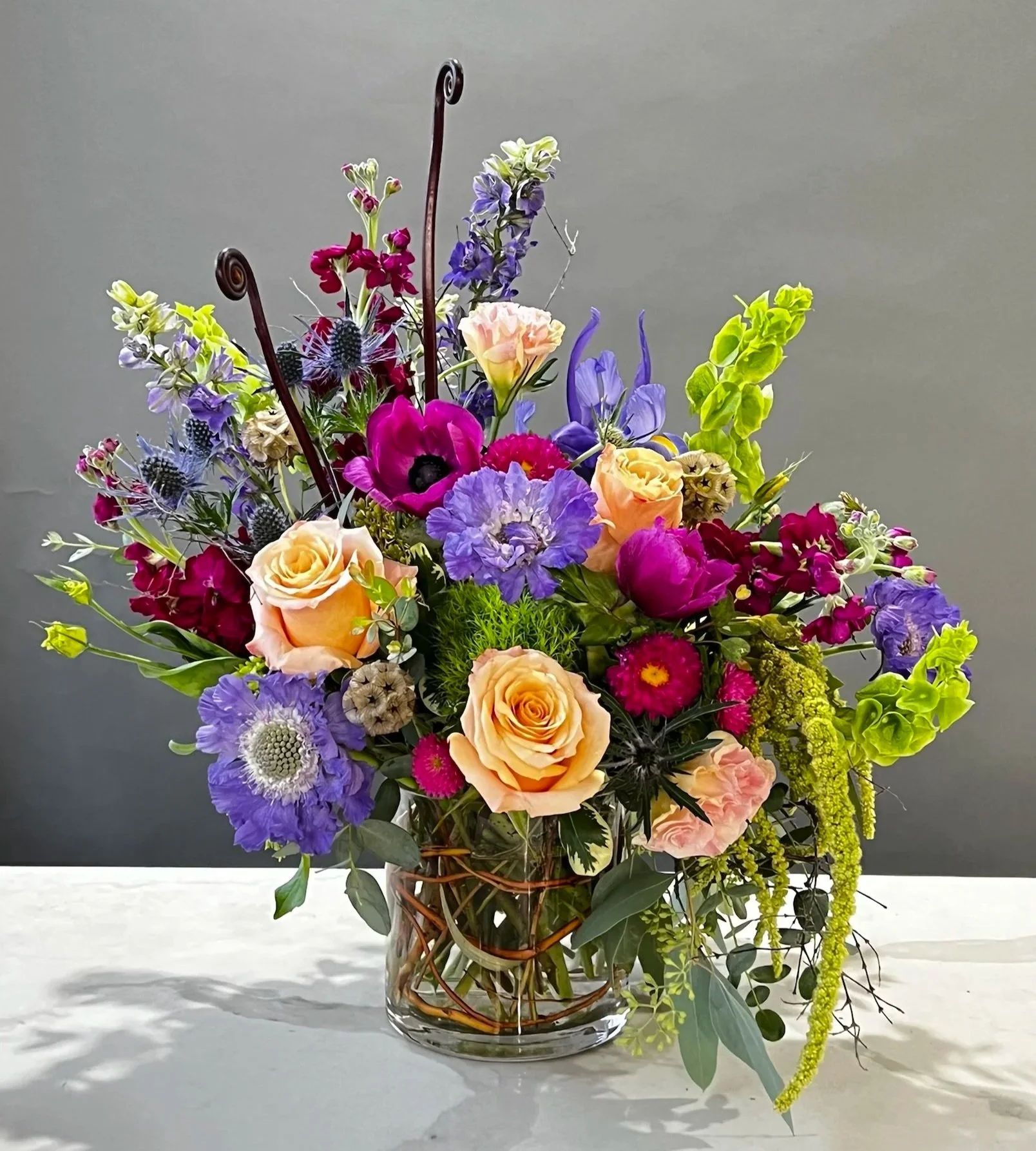 A colorful bouquet of flowers in a glass vase on a white surface against a gray background, including roses, lisianthus, delphiniums, and various greenery.