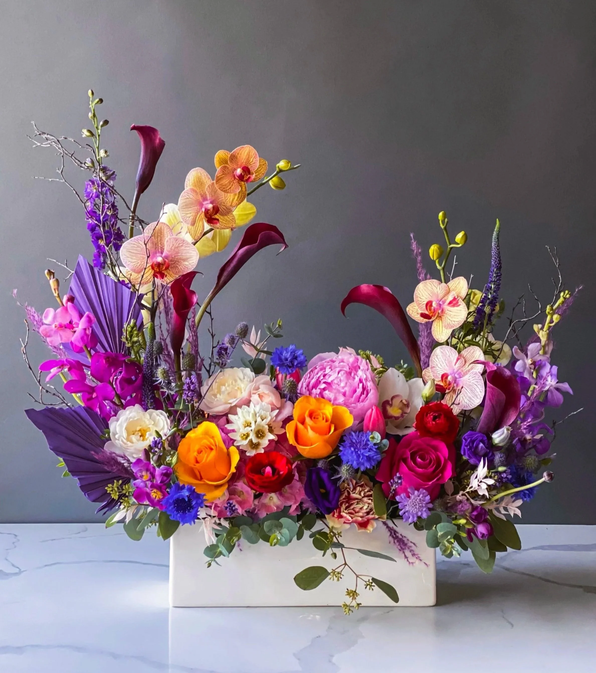 A colorful flower arrangement in a white square vase, featuring orchids, roses, carnations, and other flowers with purple, pink, yellow, and orange blooms, set against a plain gray background.