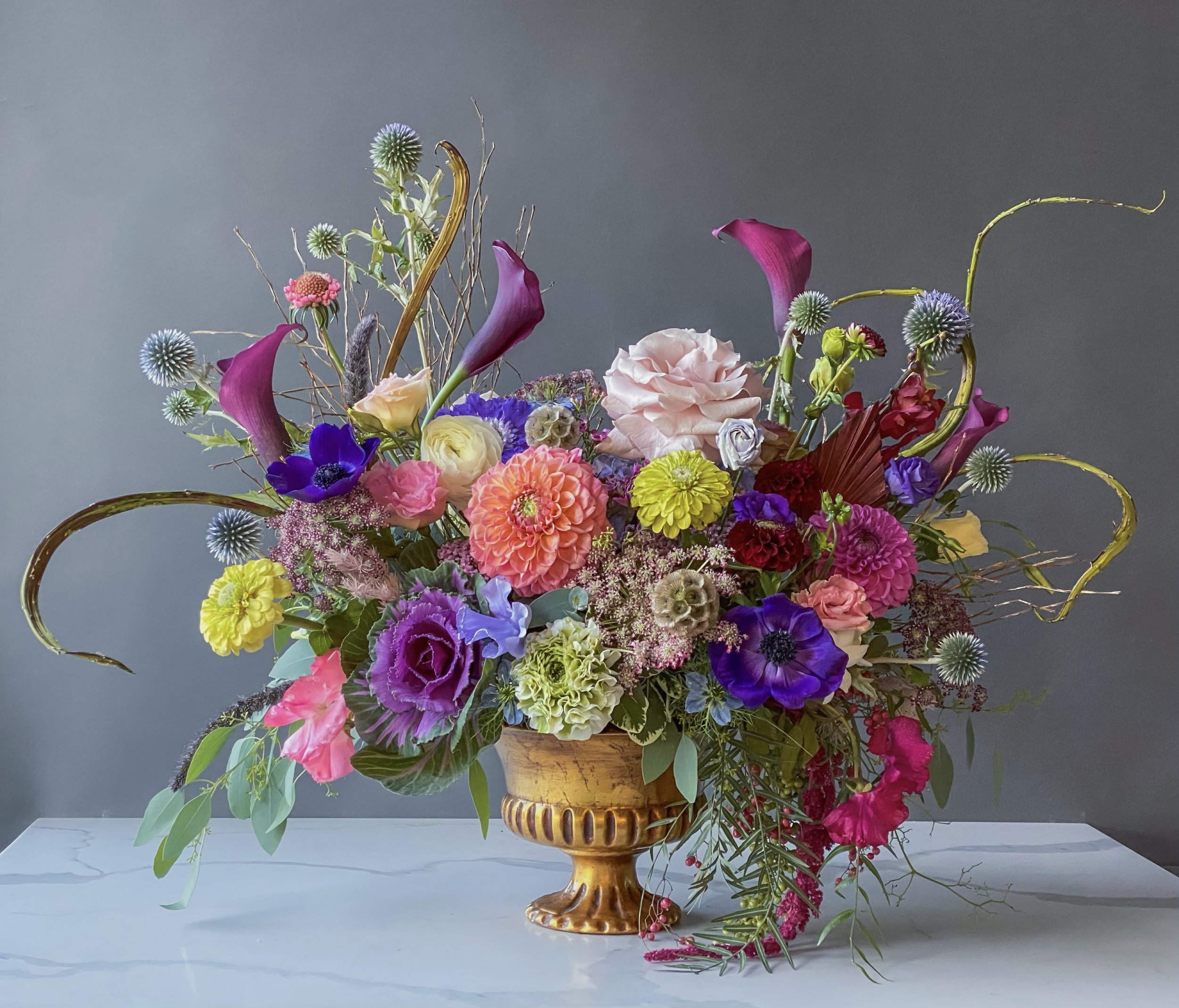 Colorful flower arrangement in a golden vase with pink, purple, yellow, white, and blue flowers, positioned against a dark gray background.