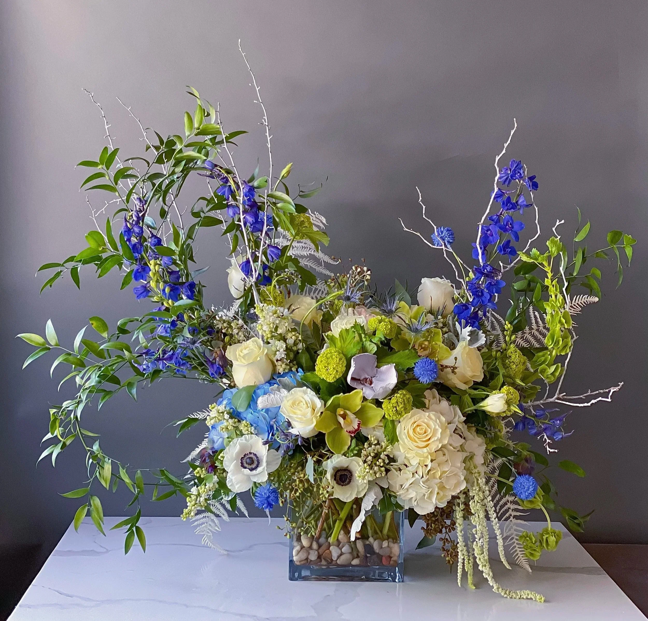 A colorful floral arrangement with white, yellow, blue, and green flowers and foliage in a square glass vase filled with small stones, set on a white surface against a gray background.