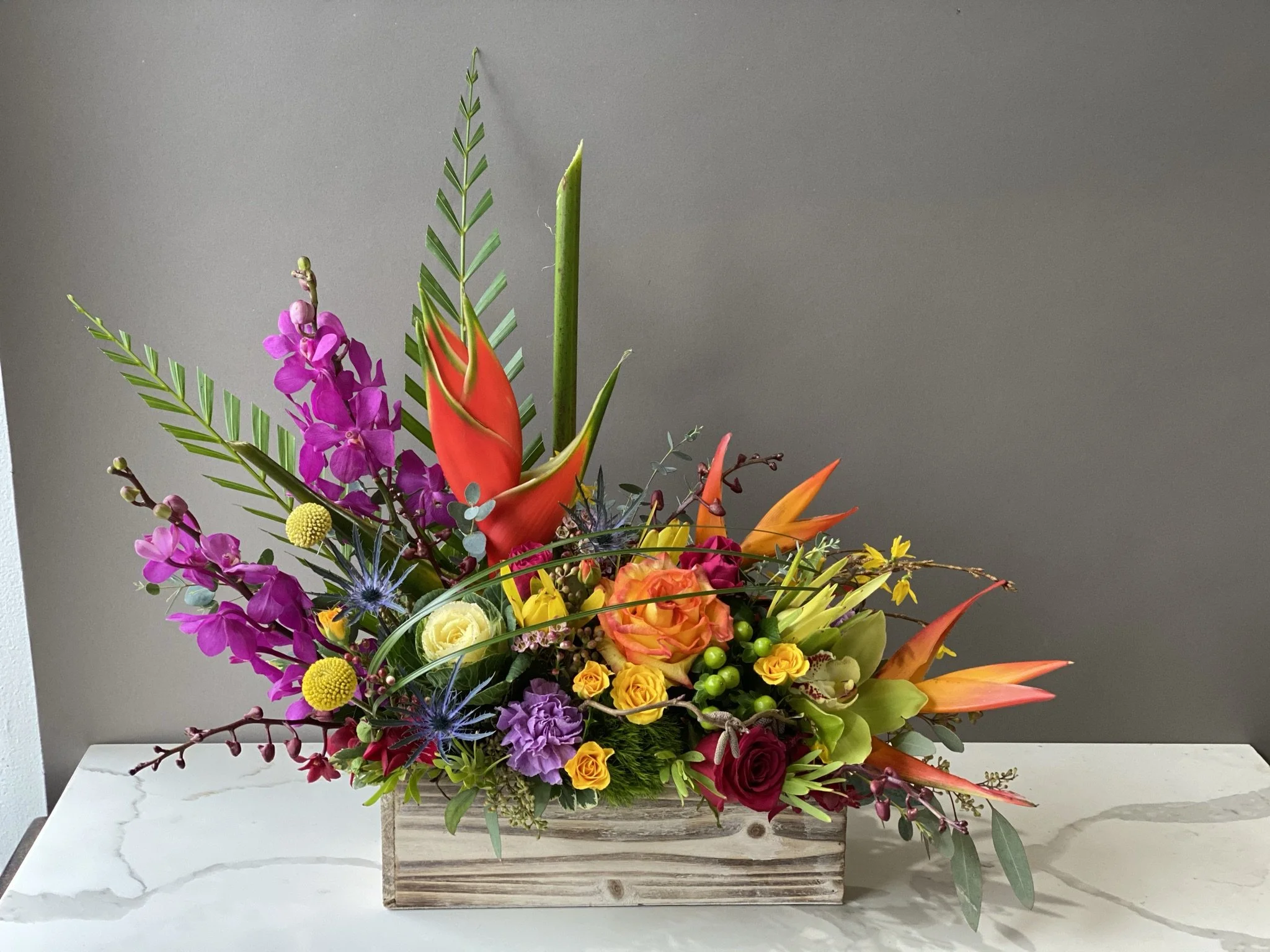 Colorful flower arrangement with pink, yellow, orange, purple, and red flowers in a wooden box on a white marble surface against a gray wall.