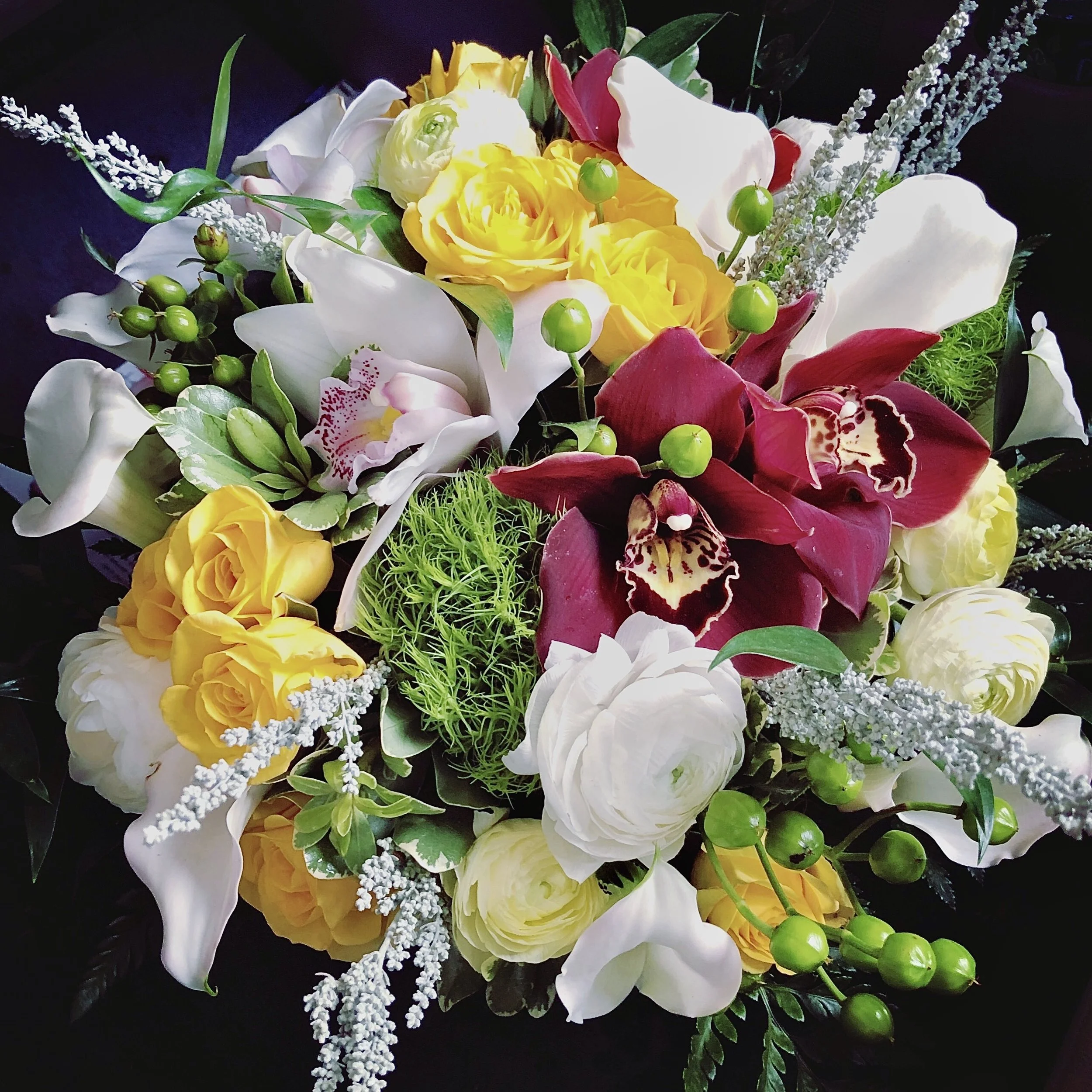 A bouquet of mixed flowers including white lilies, yellow roses, pink calla lilies, and purple orchids, accented with green foliage and small green berries.