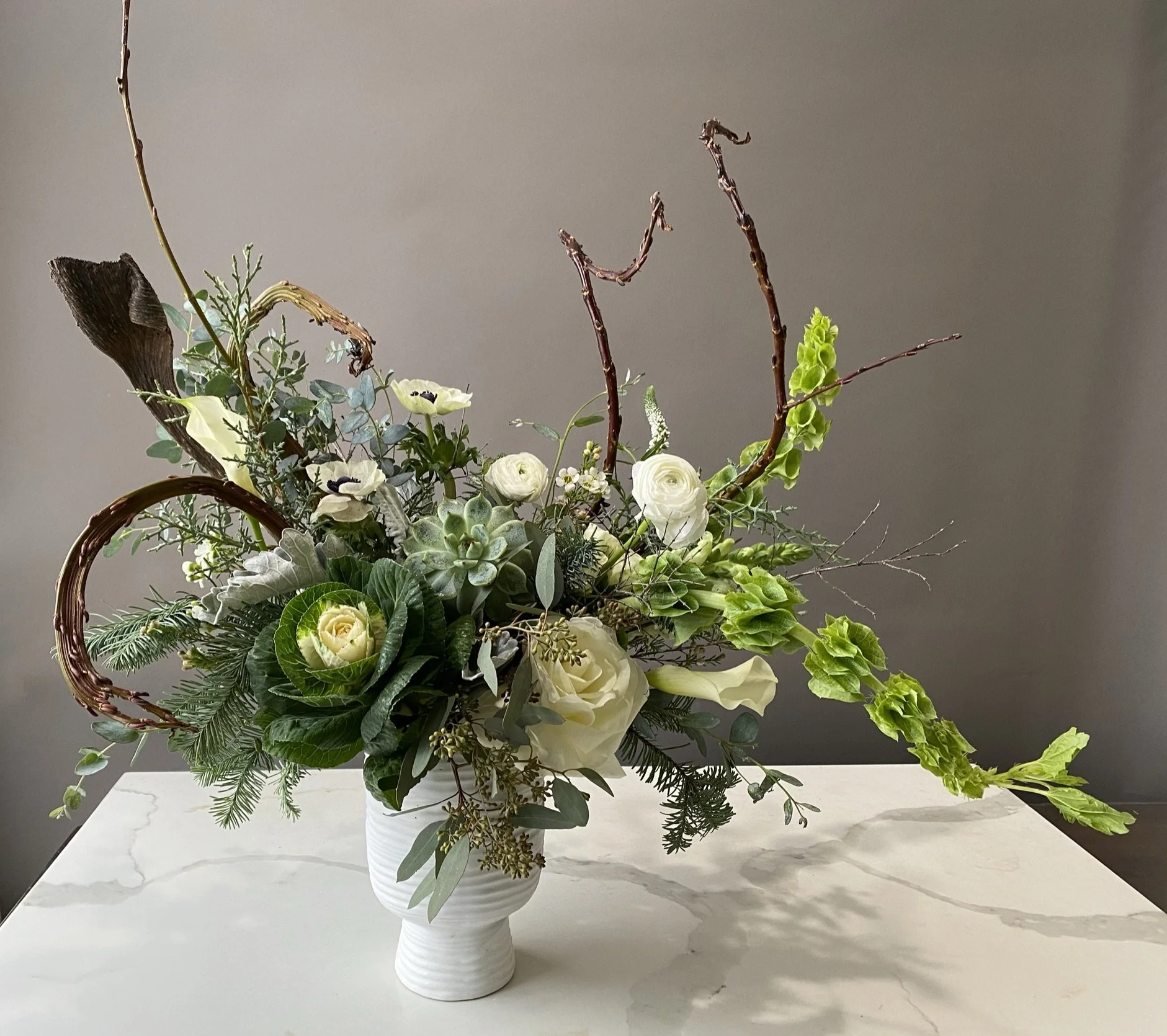 A floral arrangement in a white ceramic vase featuring white roses, various green leaves, and branches, placed on a marble surface against a plain gray wall background.