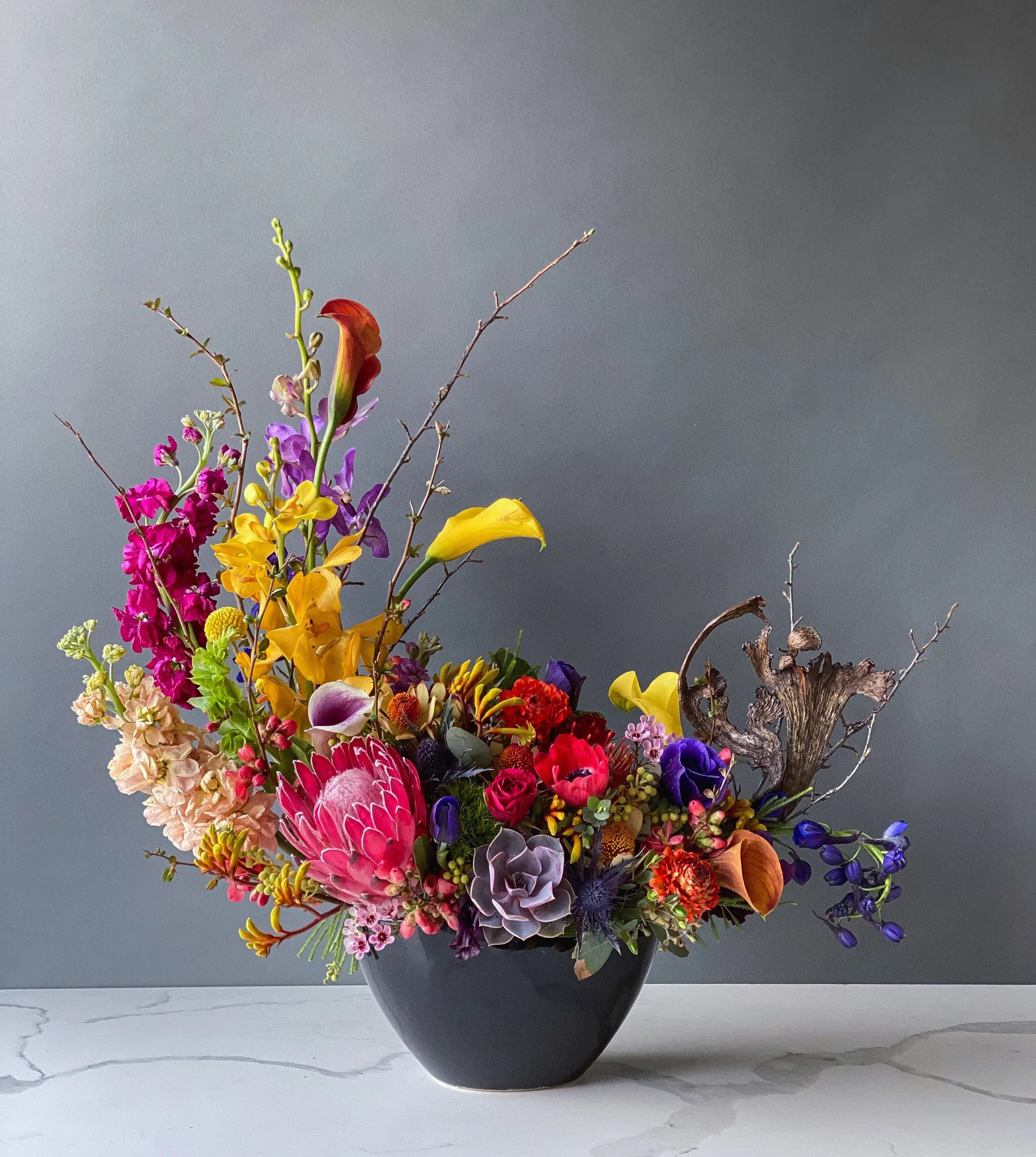 A colorful flower arrangement in a black vase on a white marble surface, with a plain gray wall in the background.