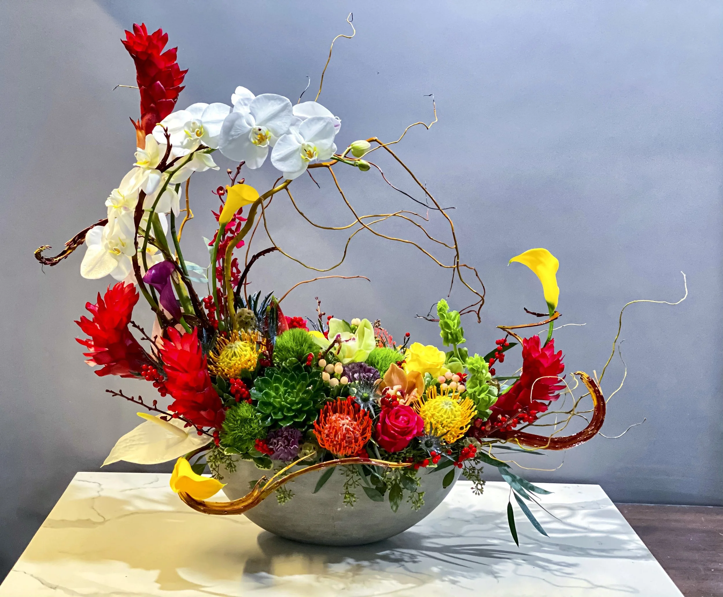 Colorful floral arrangement with white orchids, red, yellow, purple, and green flowers in a large white bowl on a marble surface against a gray background.