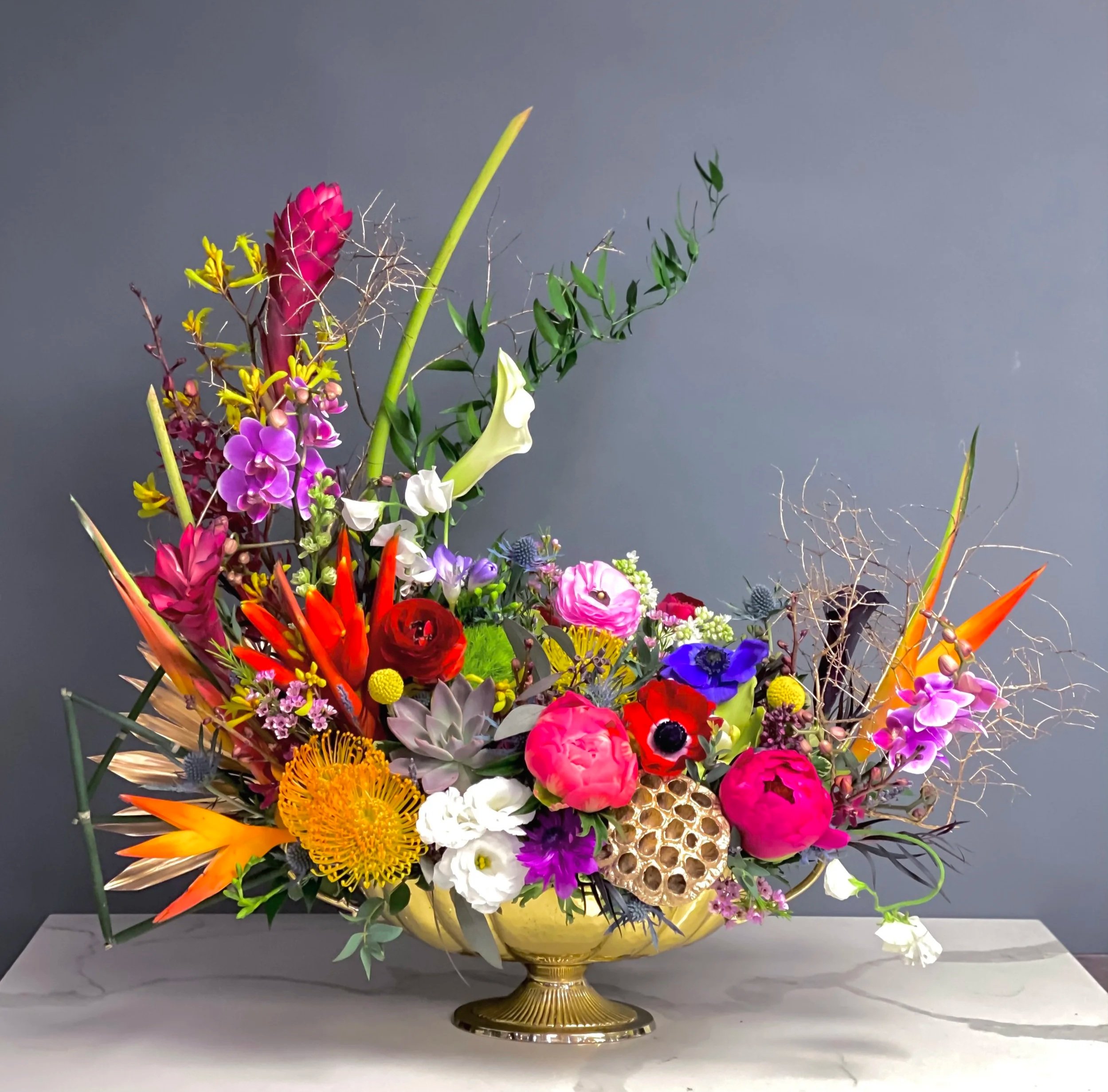 A colorful floral arrangement in a gold vase featuring various flowers including purple orchids, red roses, orange flowers, white lilies, and green leafy accents.