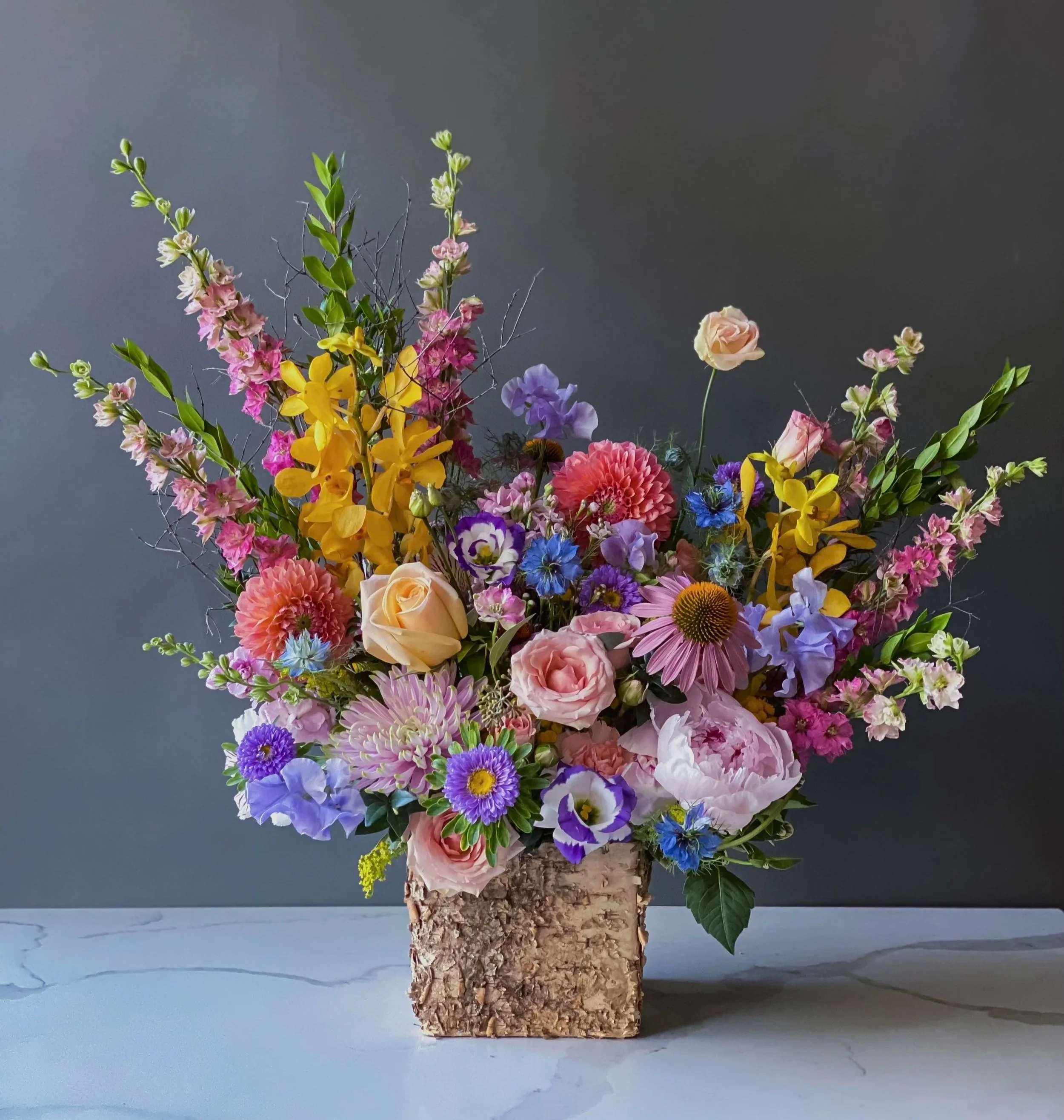 Colorful flower arrangement in a cork vase, featuring pink roses, yellow and purple flowers, green leaves, and delicate branches, placed on a light-colored surface against a gray background.