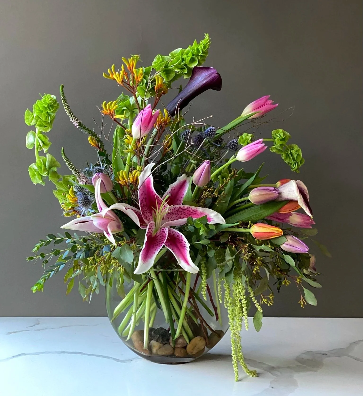 A large flower arrangement in a clear glass bowl containing lilies, calla lilies, thistles, greenery, and other assorted flowers against a gray background.