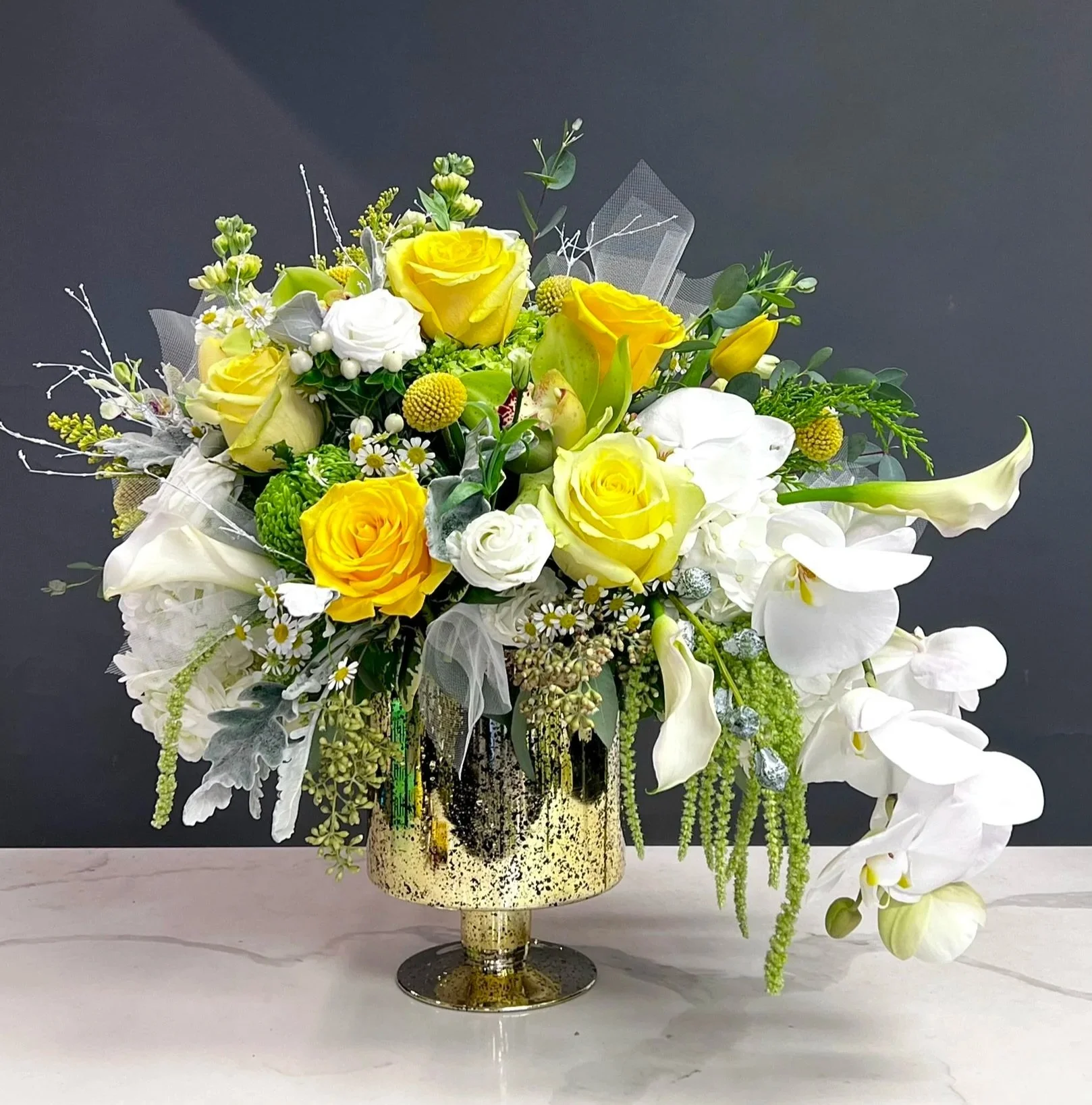 A floral arrangement with yellow roses, white roses, white orchids, green foliage, and other small flowers in a decorative gold and black vase