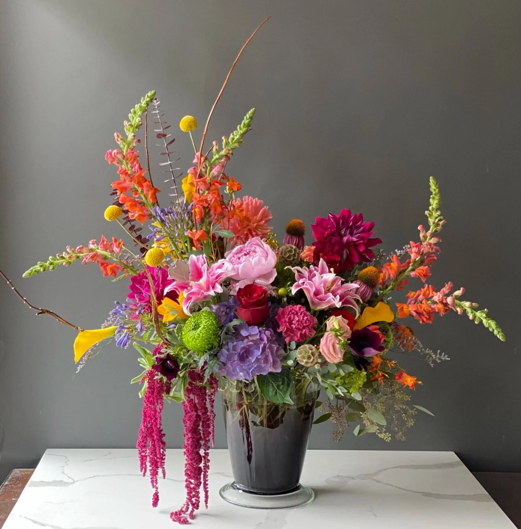 A colorful flower arrangement in a tall, dark, reflective vase on a marble table against a gray background.