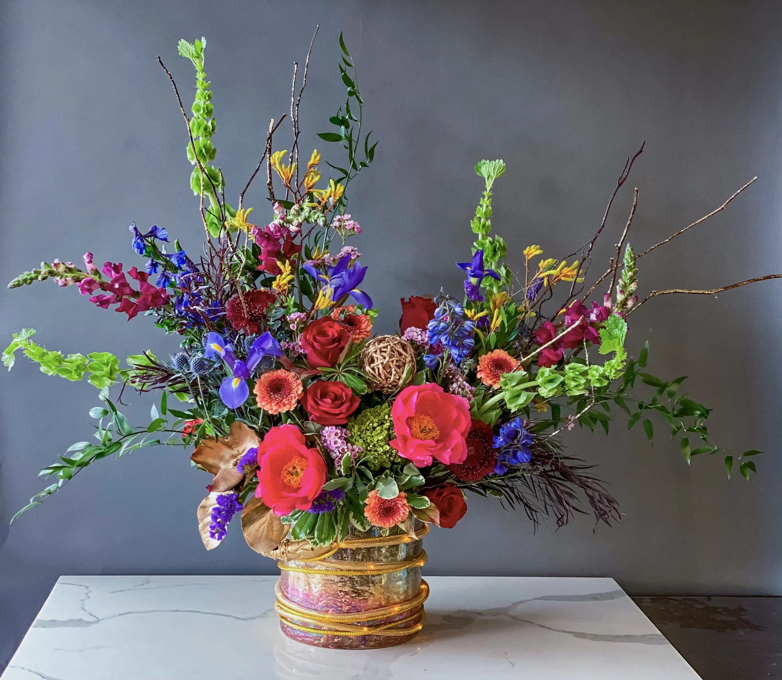A colorful bouquet of various flowers in a decorative gold and pink vase on a marble table against a plain dark background.