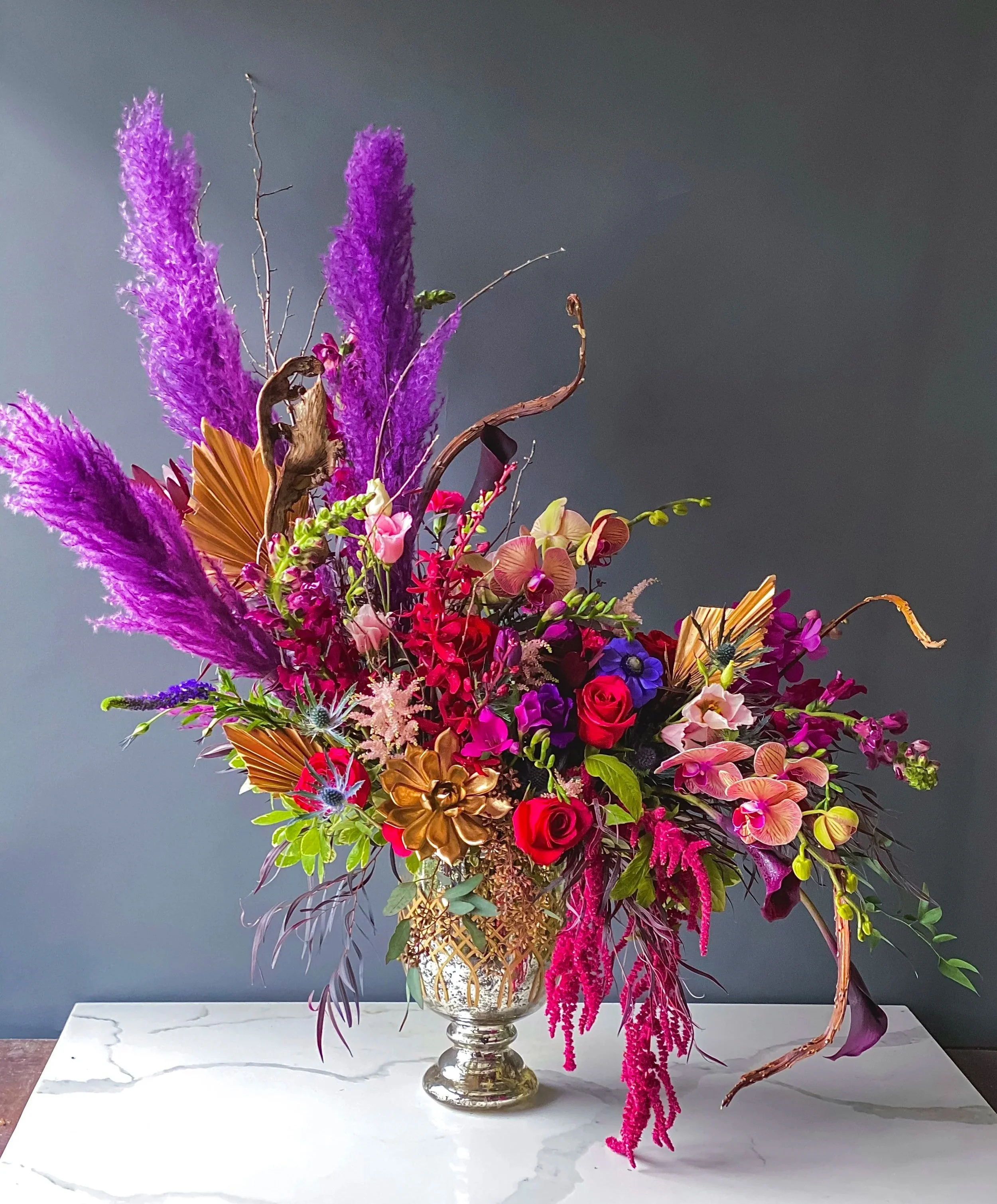 Colorful flower arrangement featuring purple, pink, yellow, and red flowers with various textures in a decorative vase on a white marble table against a dark gray background.