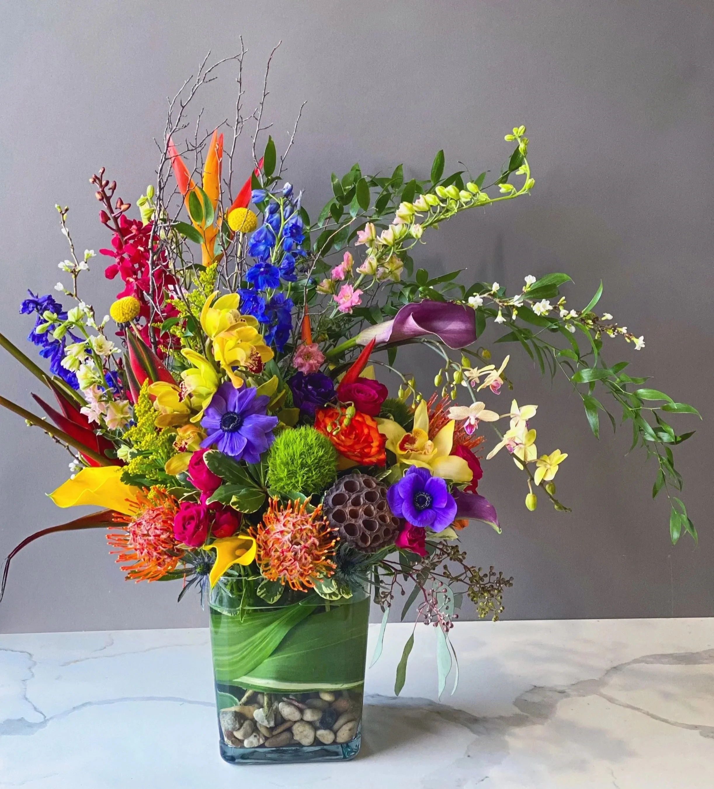 Colorful flower arrangement in a glass vase filled with decorative stones, set on a white marble surface against a gray background.