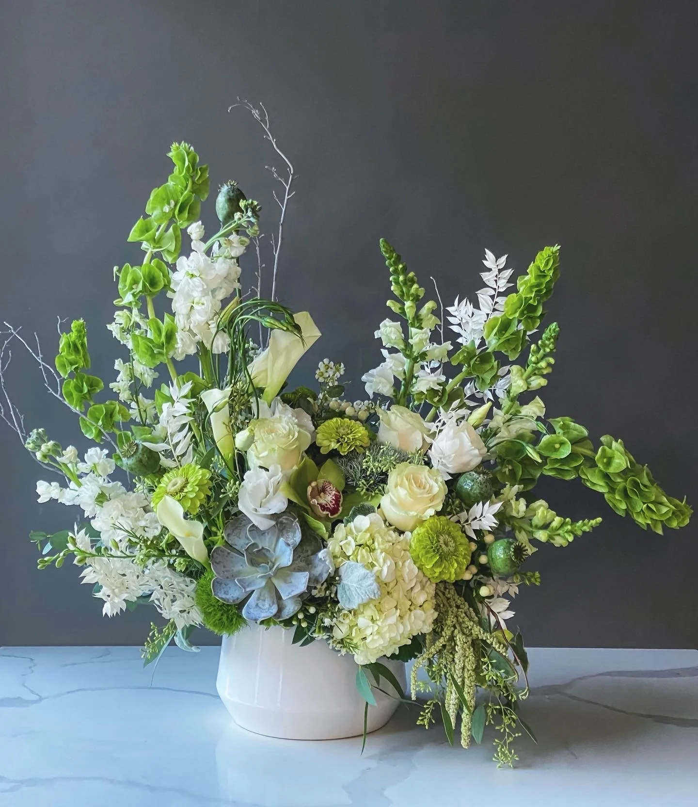 A floral arrangement in a white ceramic vase featuring white and green flowers including roses, hydrangeas, and succulents against a dark background.