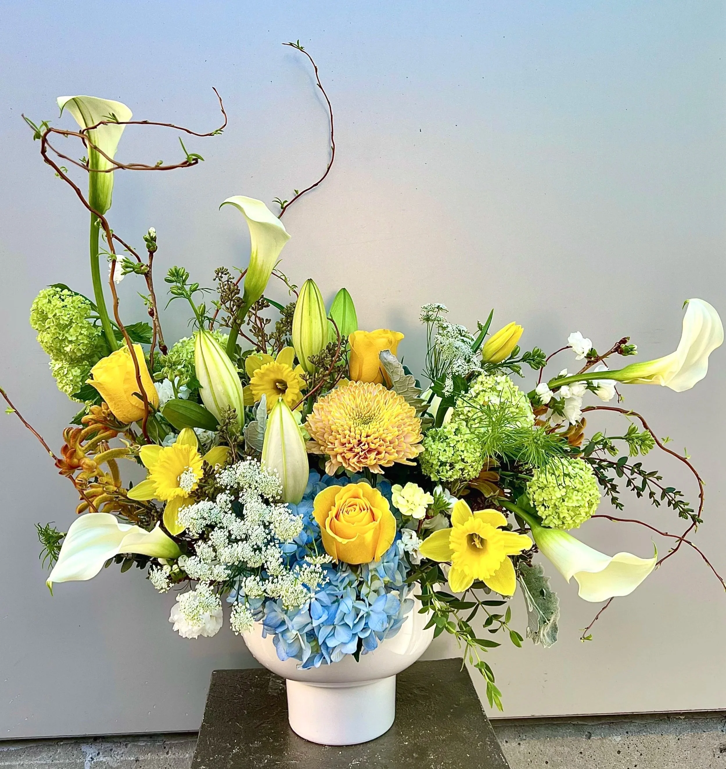 A floral arrangement in a white vase containing white lilies, yellow roses, purple and yellow flowers, white and green hydrangeas, small white flowers, and greenery against a plain light grey background.