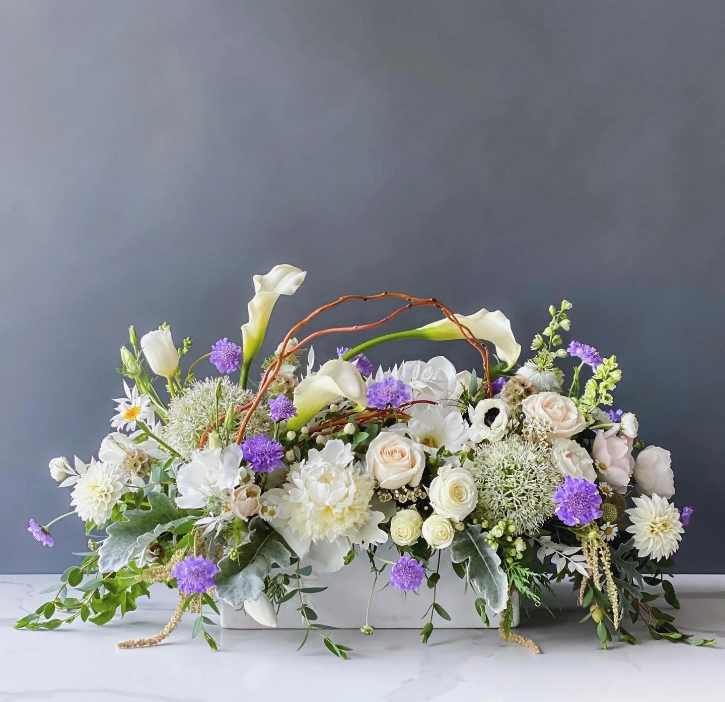 Elegant floral arrangement in a white container with white, purple, and green flowers and foliage, set against a gray background.