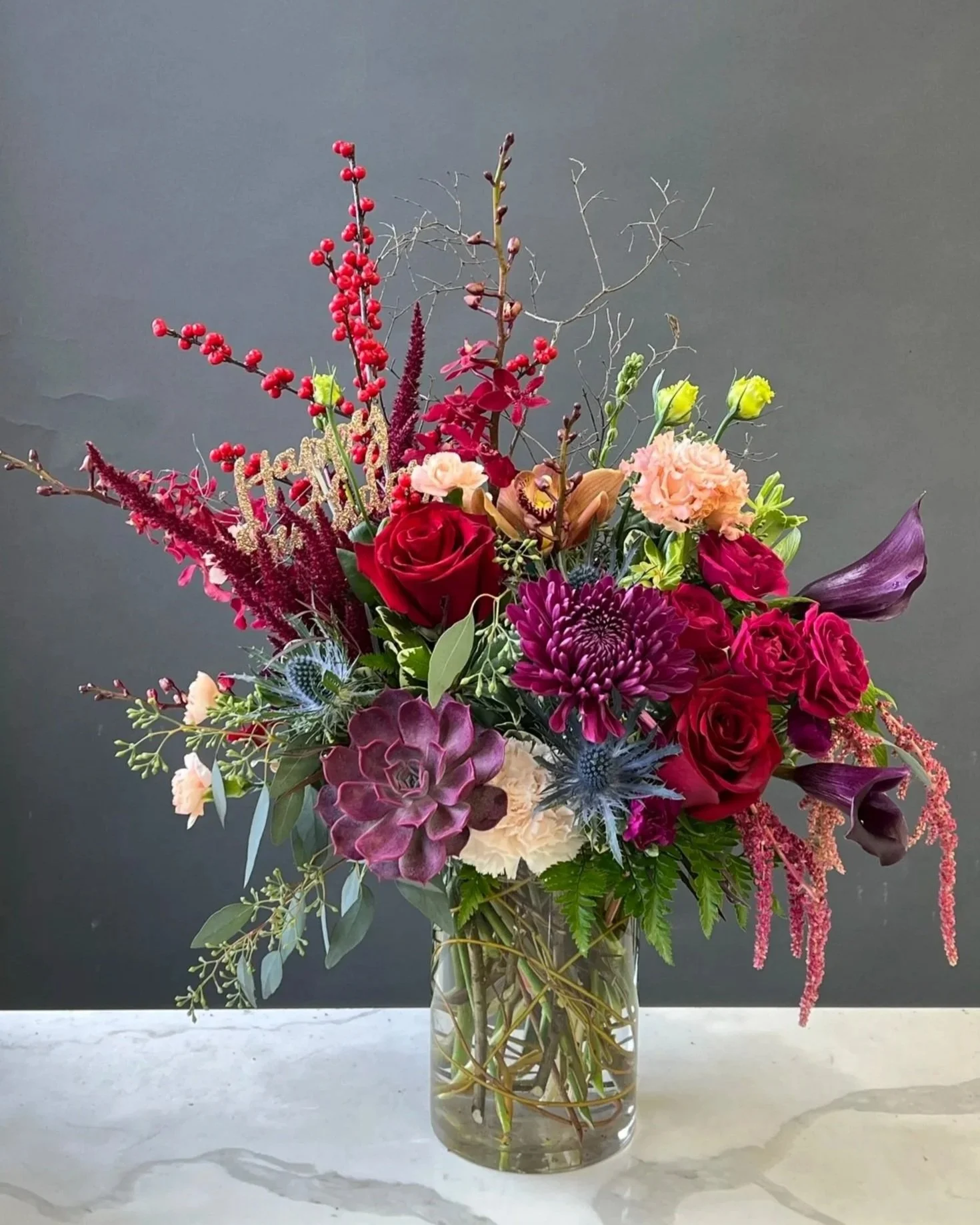 A colorful flower bouquet in a clear glass vase placed on a marble surface against a gray background. The bouquet includes red roses, pink carnations, purple calla lilies, blue thistles, and various greenery and accents.