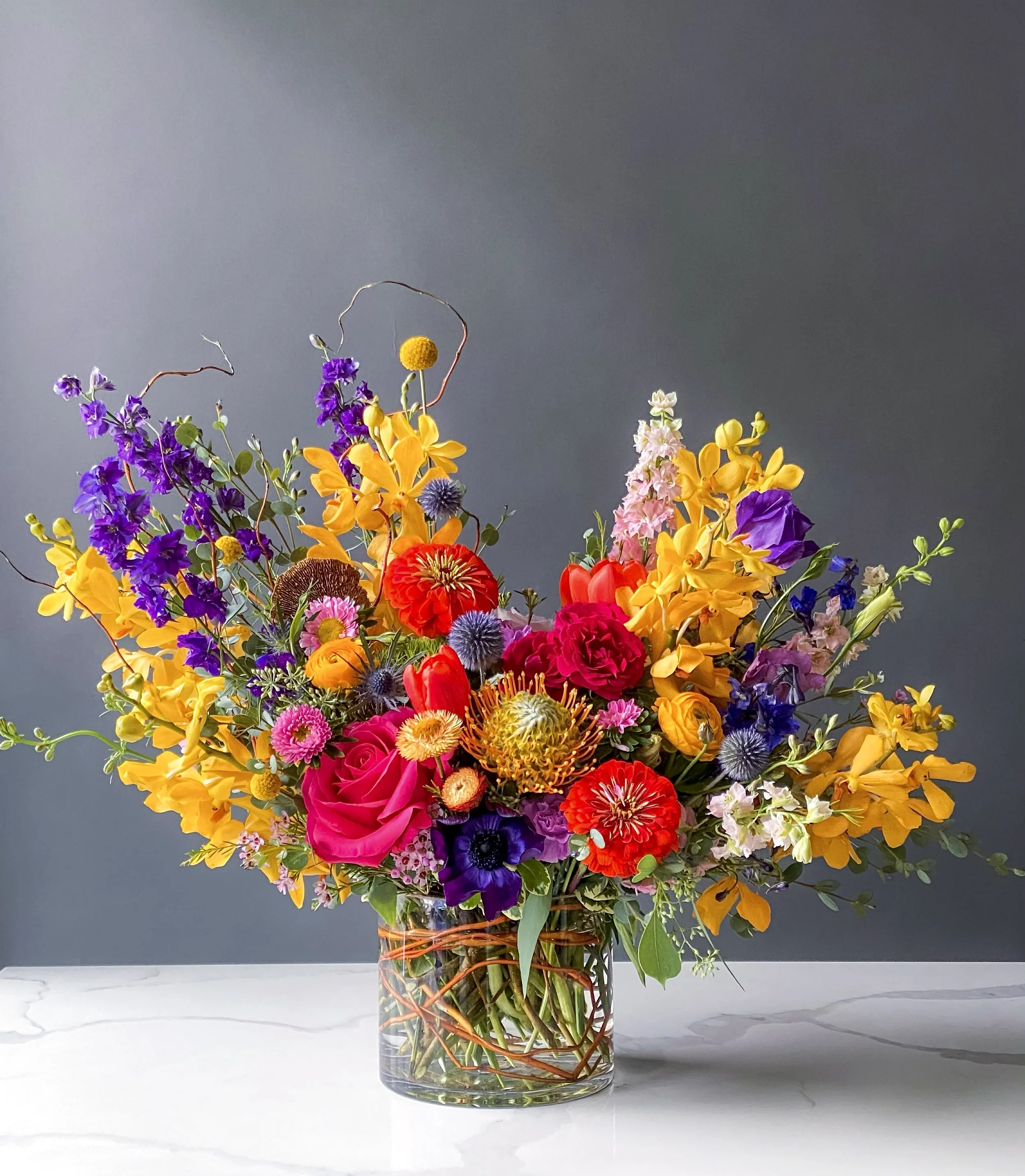 A large bouquet of colorful flowers in a clear glass vase on a white marble surface against a gray background.
