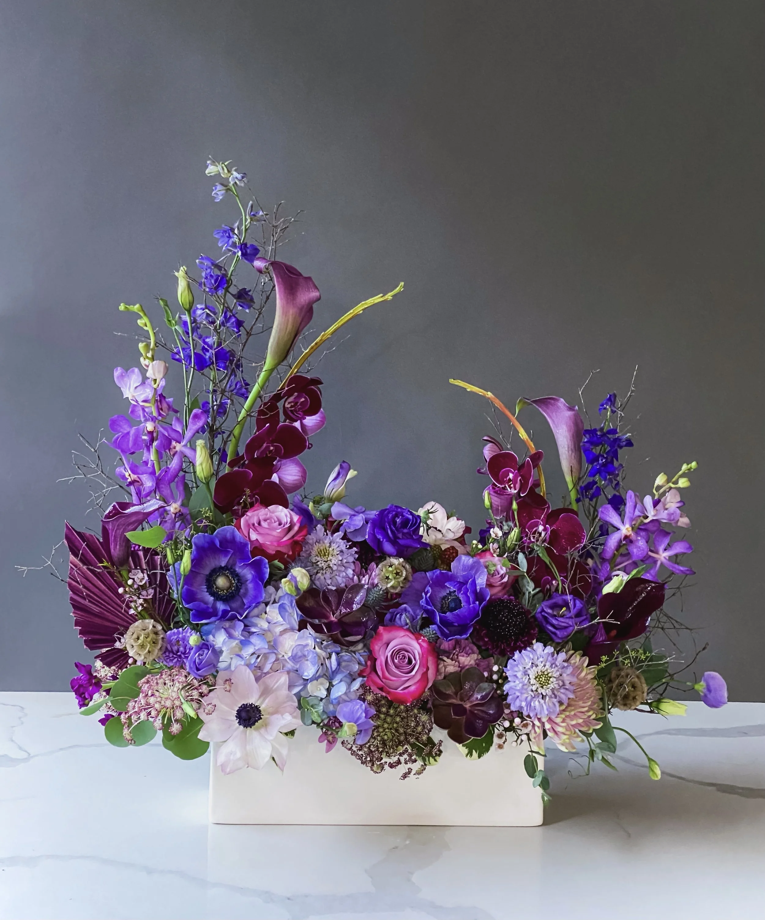 A colorful flower arrangement with purple, pink, white, and red flowers in a white rectangular vase against a dark background.