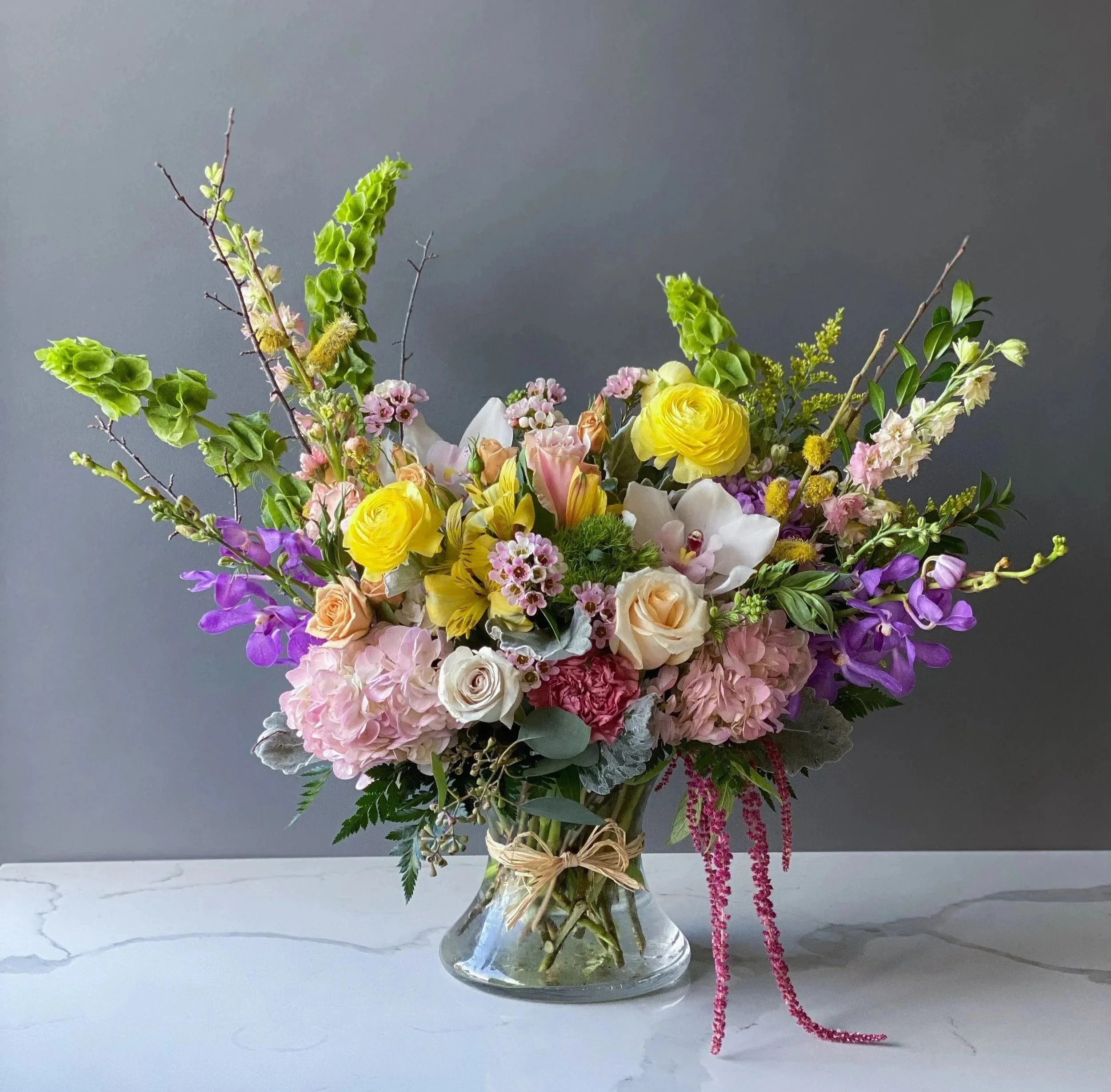 A colorful flower arrangement in a glass vase with pink, yellow, white, and purple flowers and greenery against a gray background.