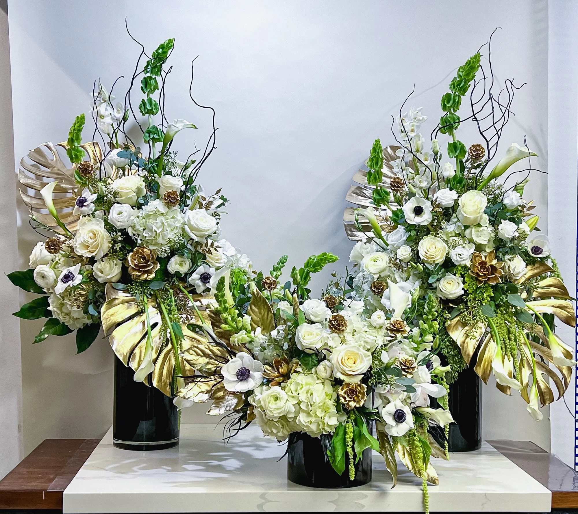 A display of two floral arrangements in black vases, consisting of white roses, calla lilies, anemones, and various greenery, arranged on a white table against a plain wall.