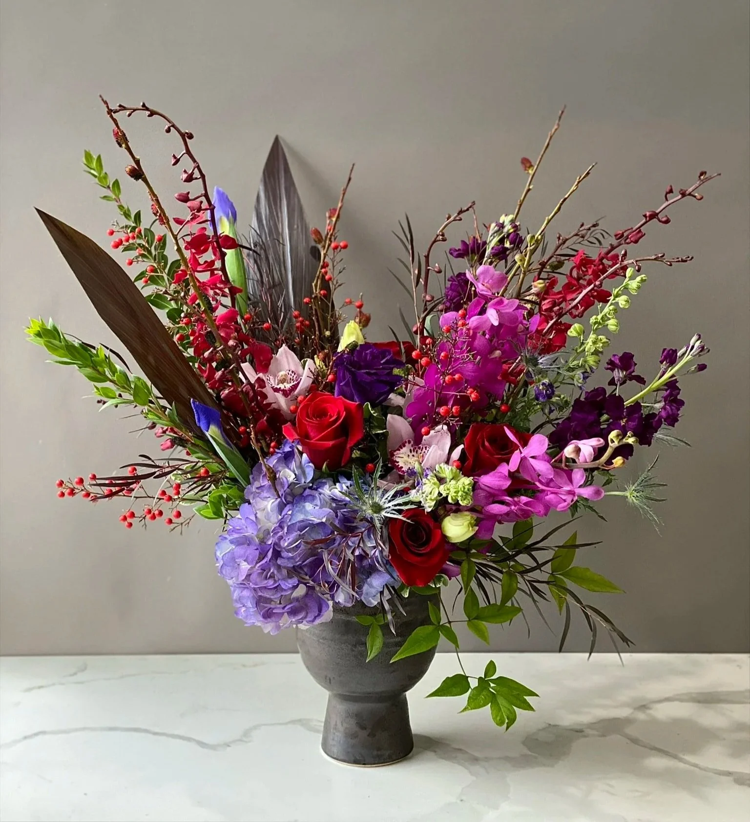 A tall, gray ceramic vase filled with a colorful bouquet of various flowers and greenery, set on a light-colored surface against a plain gray wall.
