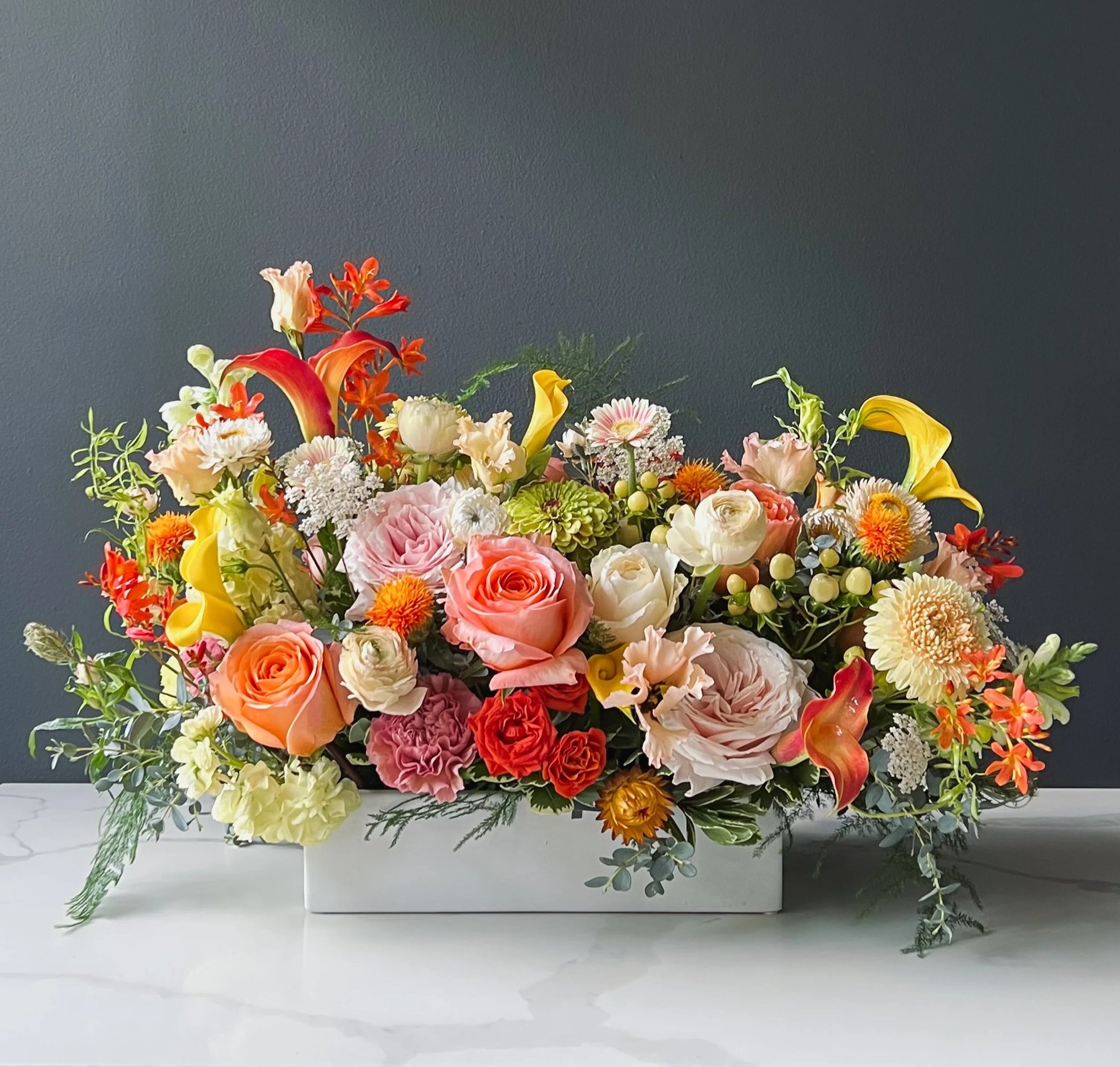 Large colorful flower arrangement with roses, lilies, and other mixed flowers placed in a white rectangular box against a dark gray wall.
