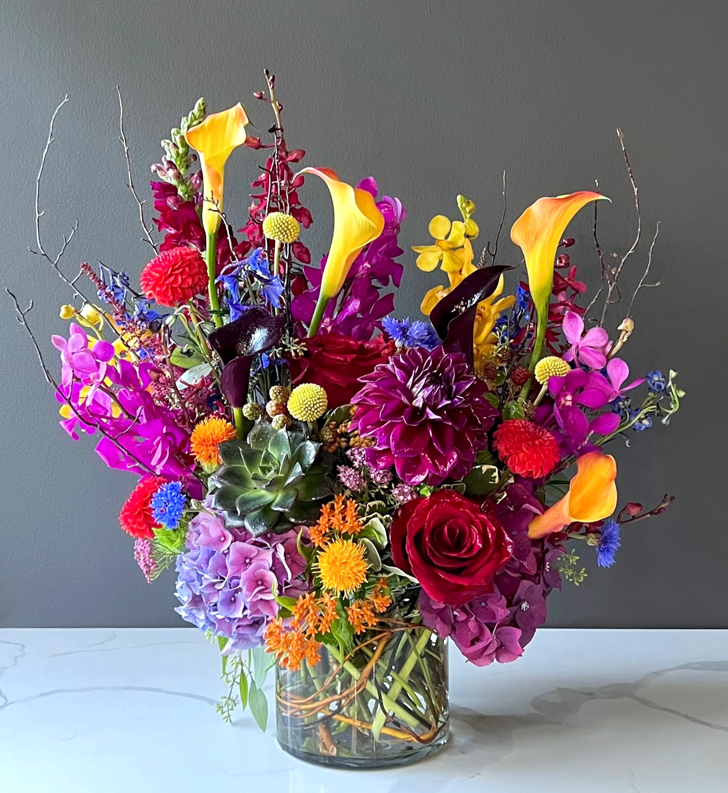 A vibrant bouquet of multicolored flowers in a clear glass vase, set against a gray backdrop. The arrangement includes yellow calla lilies, purple orchids, red dahlias, blue asters, orange chrysanthemums, and other assorted blooms and greenery.