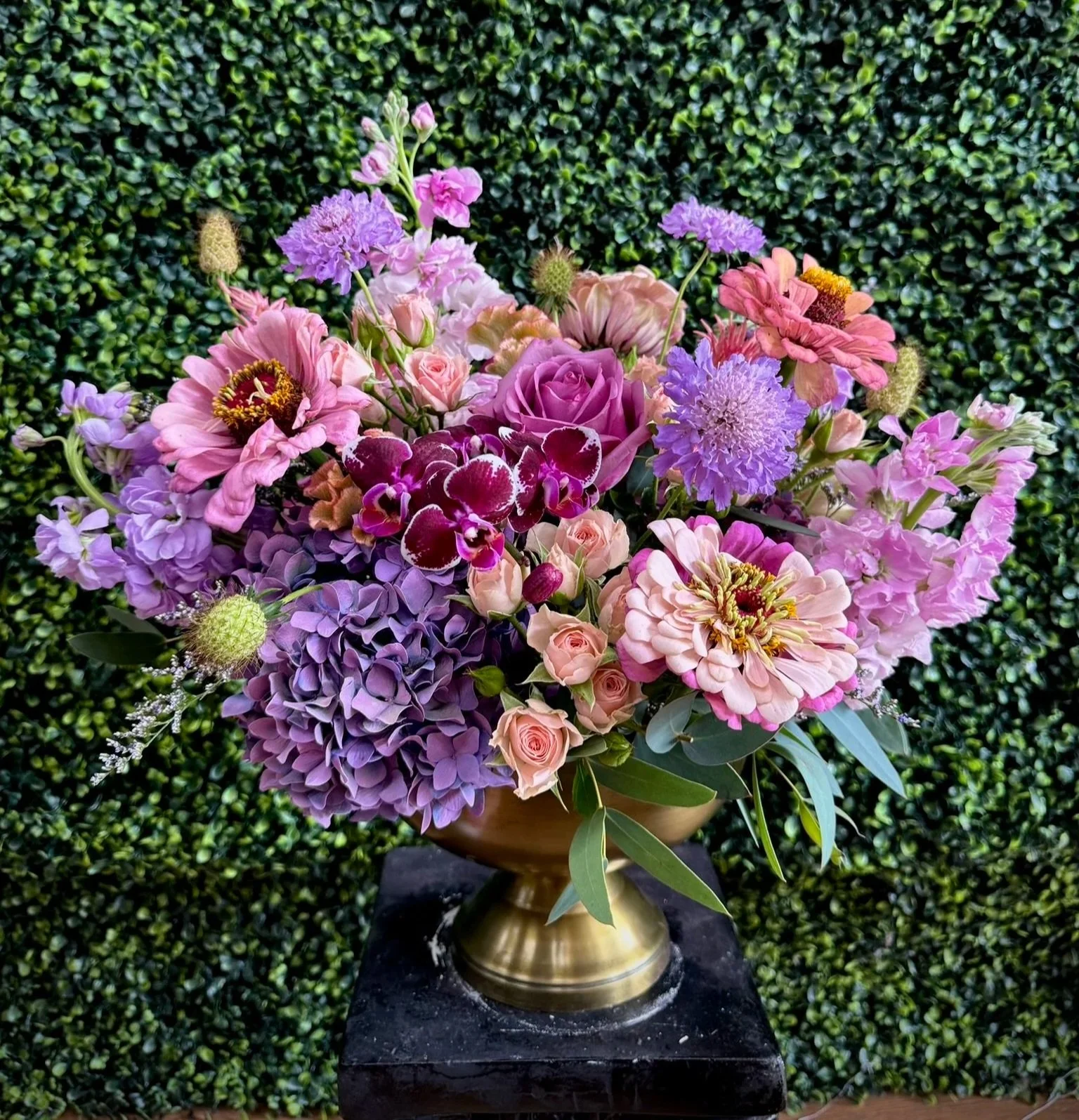 A colorful bouquet of various flowers including roses, hydrangeas, and zinnias in a bronze vase placed on a dark surface against a green leafy background.