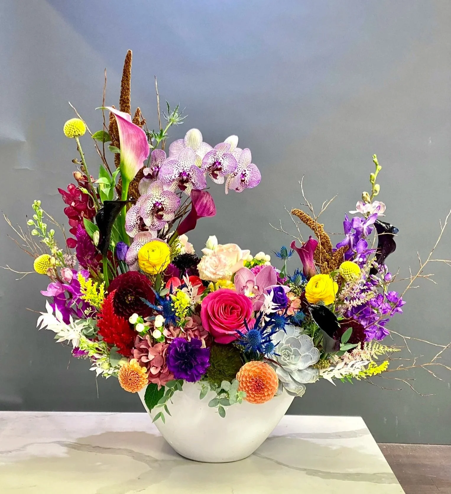 A colorful flower arrangement in a white ceramic vase on a white table against a gray background. The bouquet includes roses, orchids, dahlias, calla lilies, and other assorted flowers and greenery.