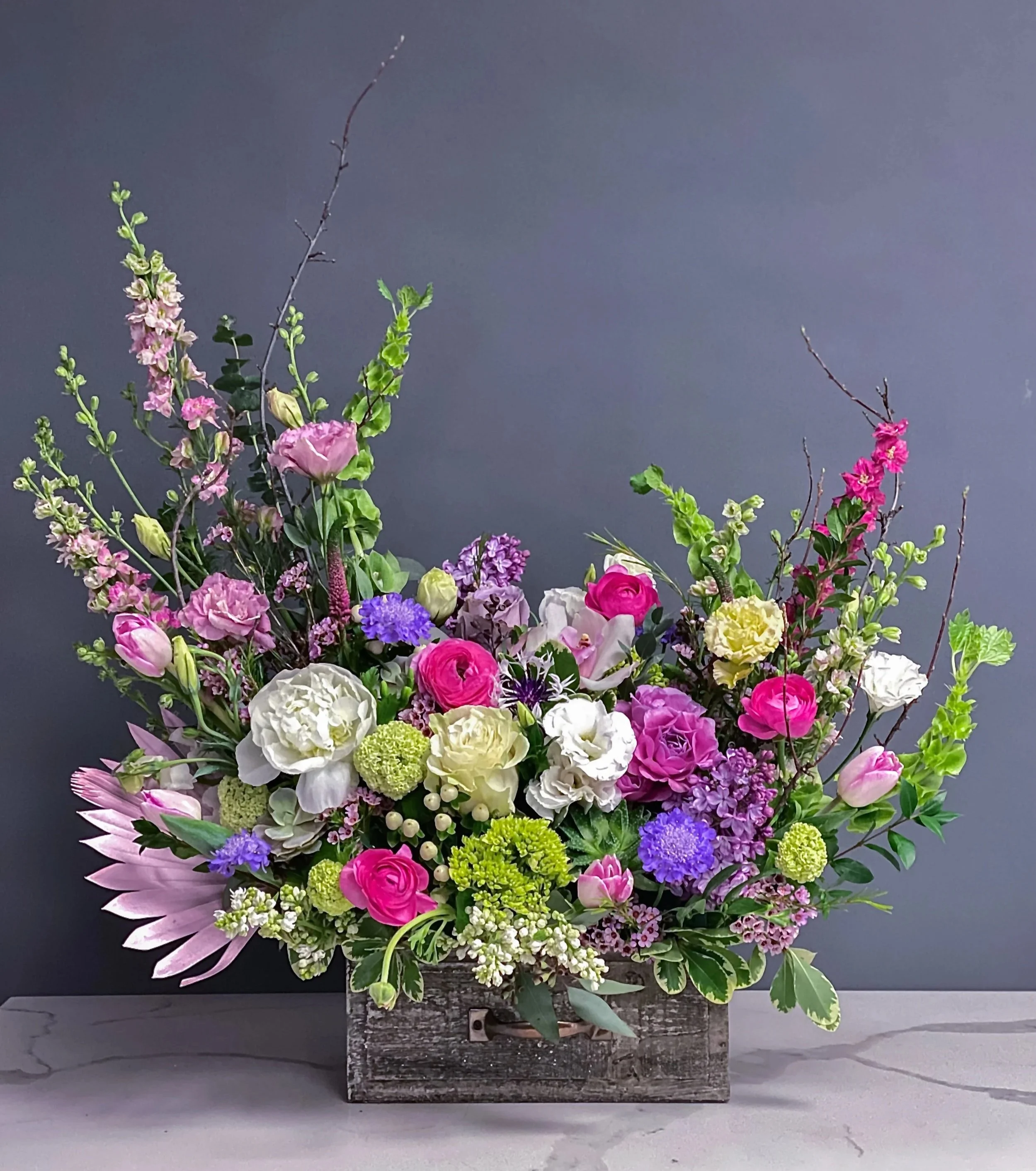A large, colorful flower arrangement in a rustic wooden box, featuring pink roses, white carnations, purple and pink snapdragons, green hydrangeas, and various greenery, set against a gray backdrop.