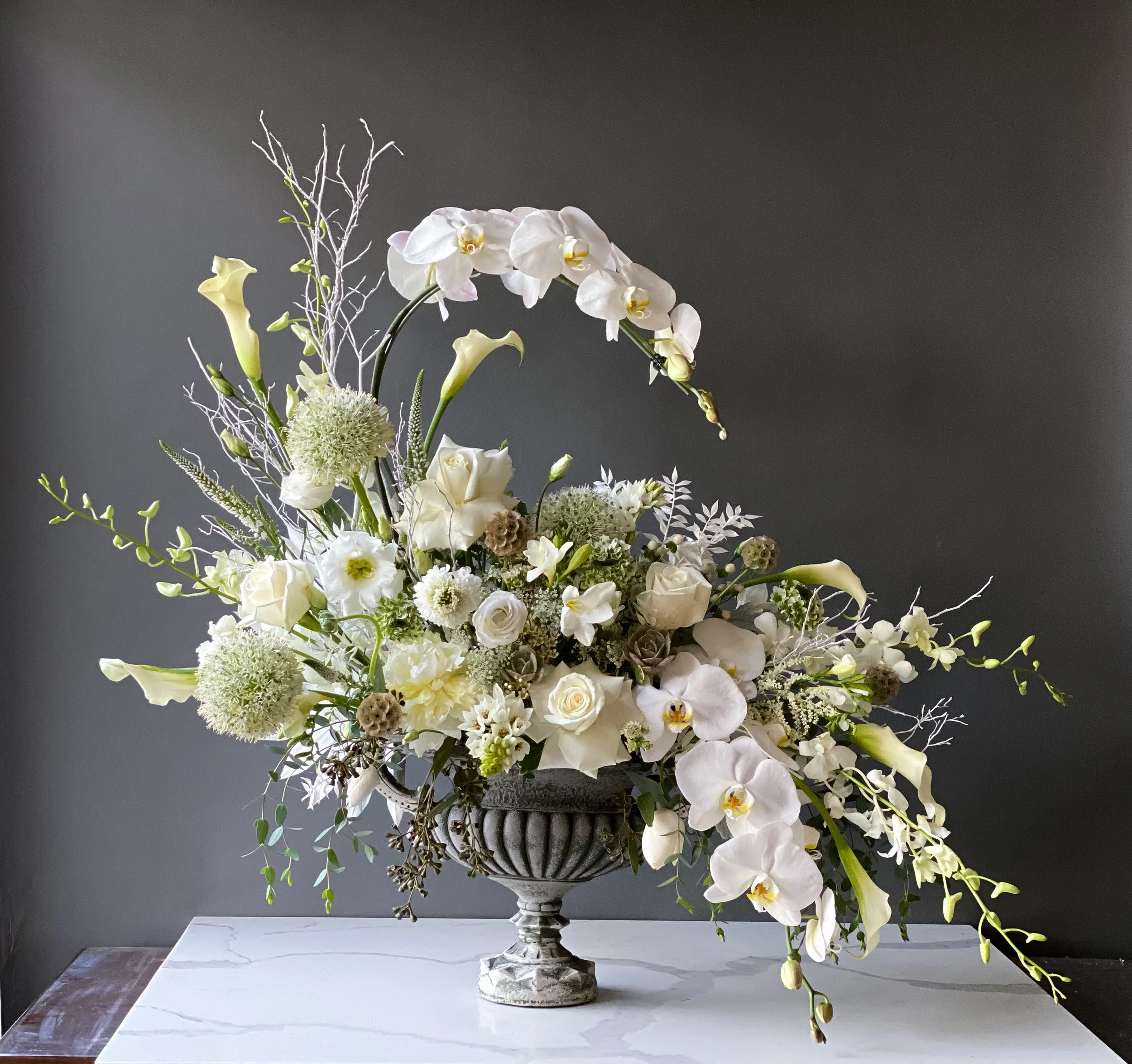 Elegant floral arrangement with white orchids, calla lilies, roses, and greenery in a decorative silver vase on a white marble table against a dark gray background.
