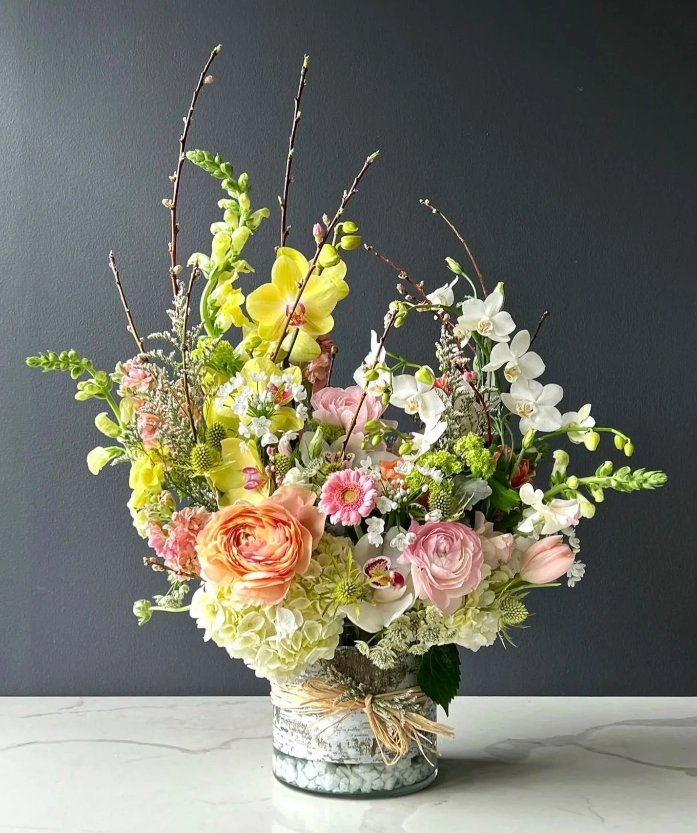A colorful flower bouquet in a white patterned vase on a white marble surface, featuring pink roses, white orchids, yellow lilies, and other assorted flowers and greenery against a dark gray background.