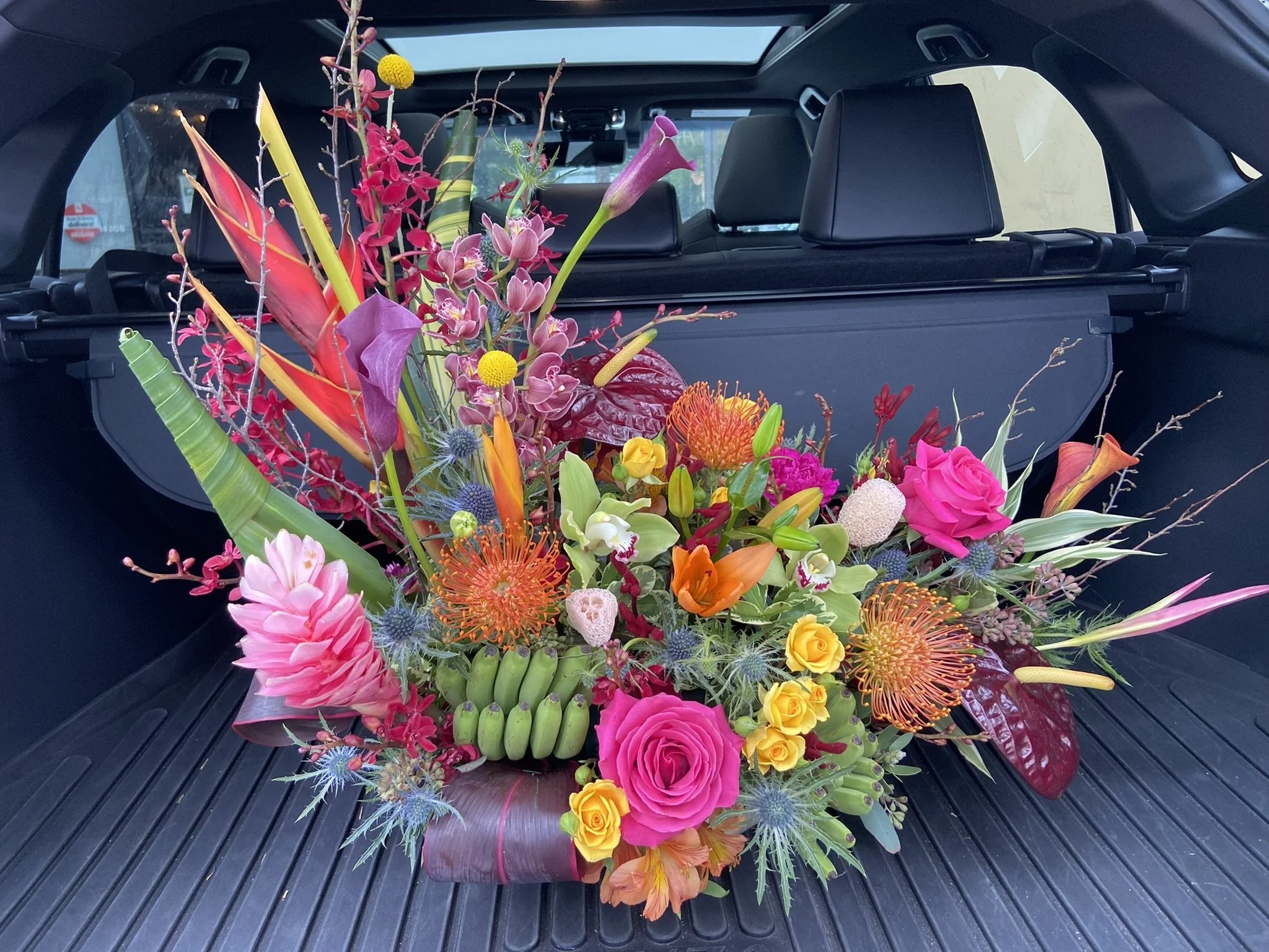 A colorful flower bouquet with pink, yellow, orange, purple, and blue flowers inside the trunk of a car with a black interior and a sunroof.
