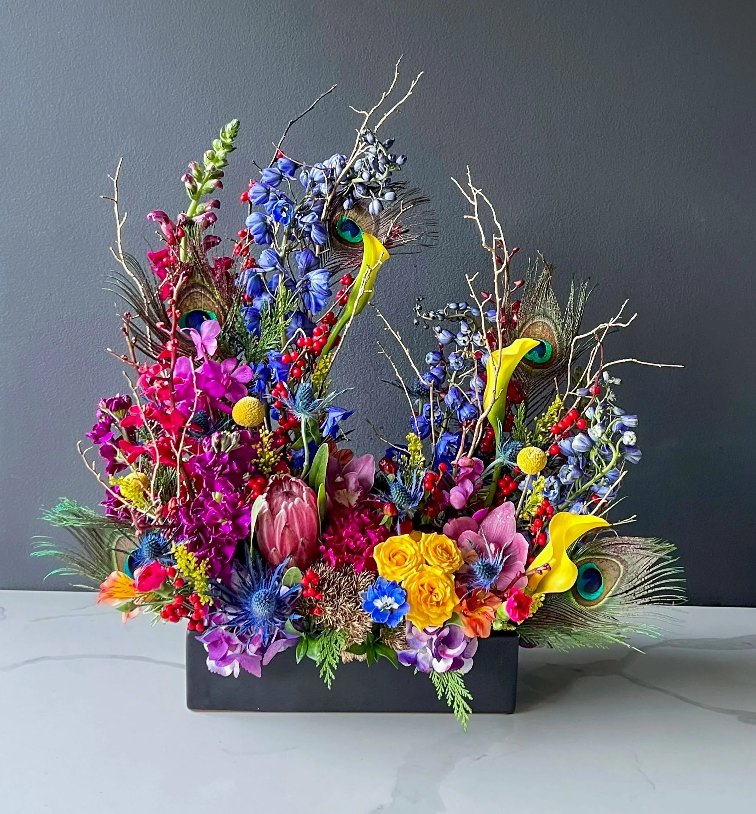 Colorful flower arrangement with peacock feathers in a black rectangular vase against a gray wall.